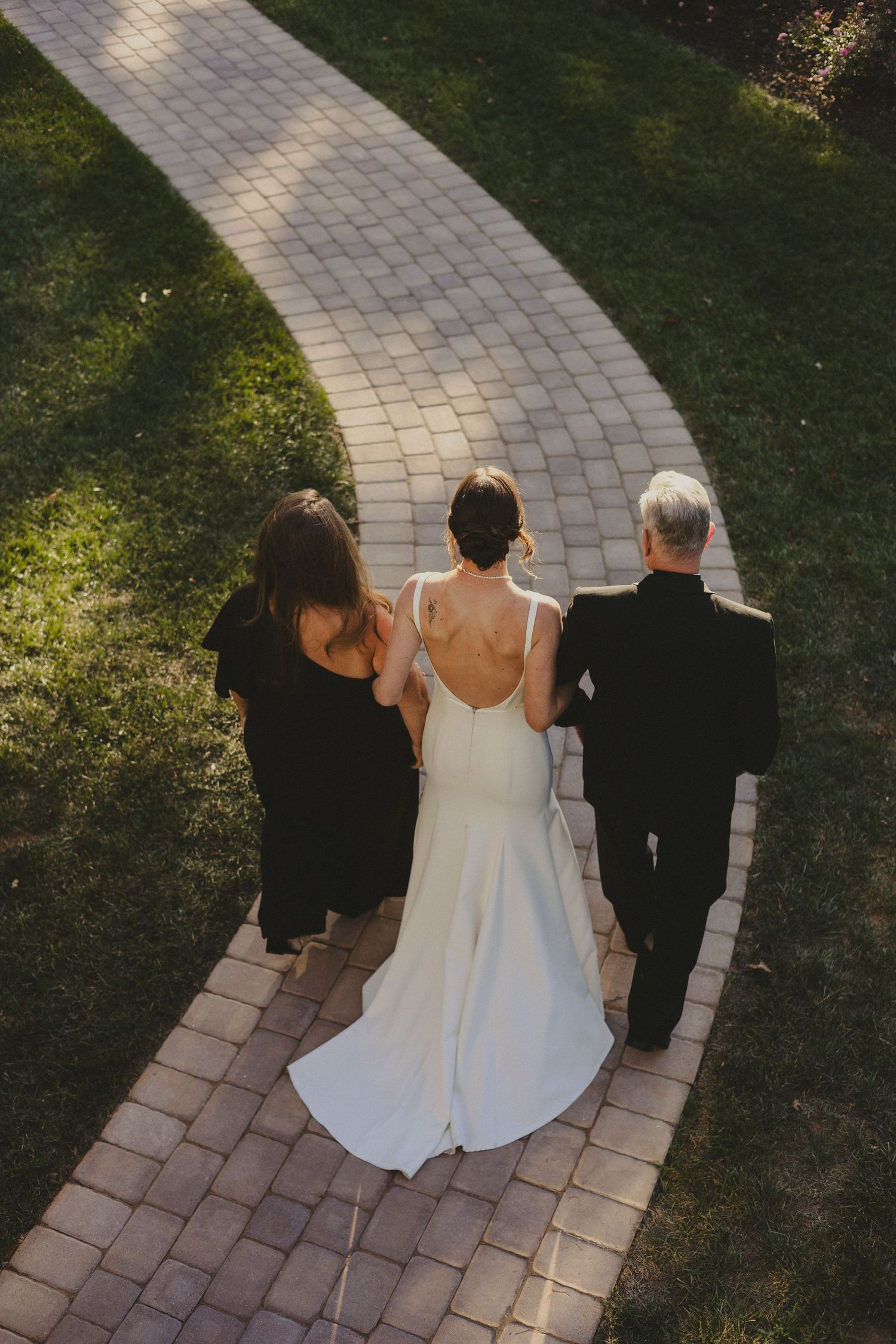 Aerial view of people walking on a curved stone pathway through a green lawn during a wedding ceremony.