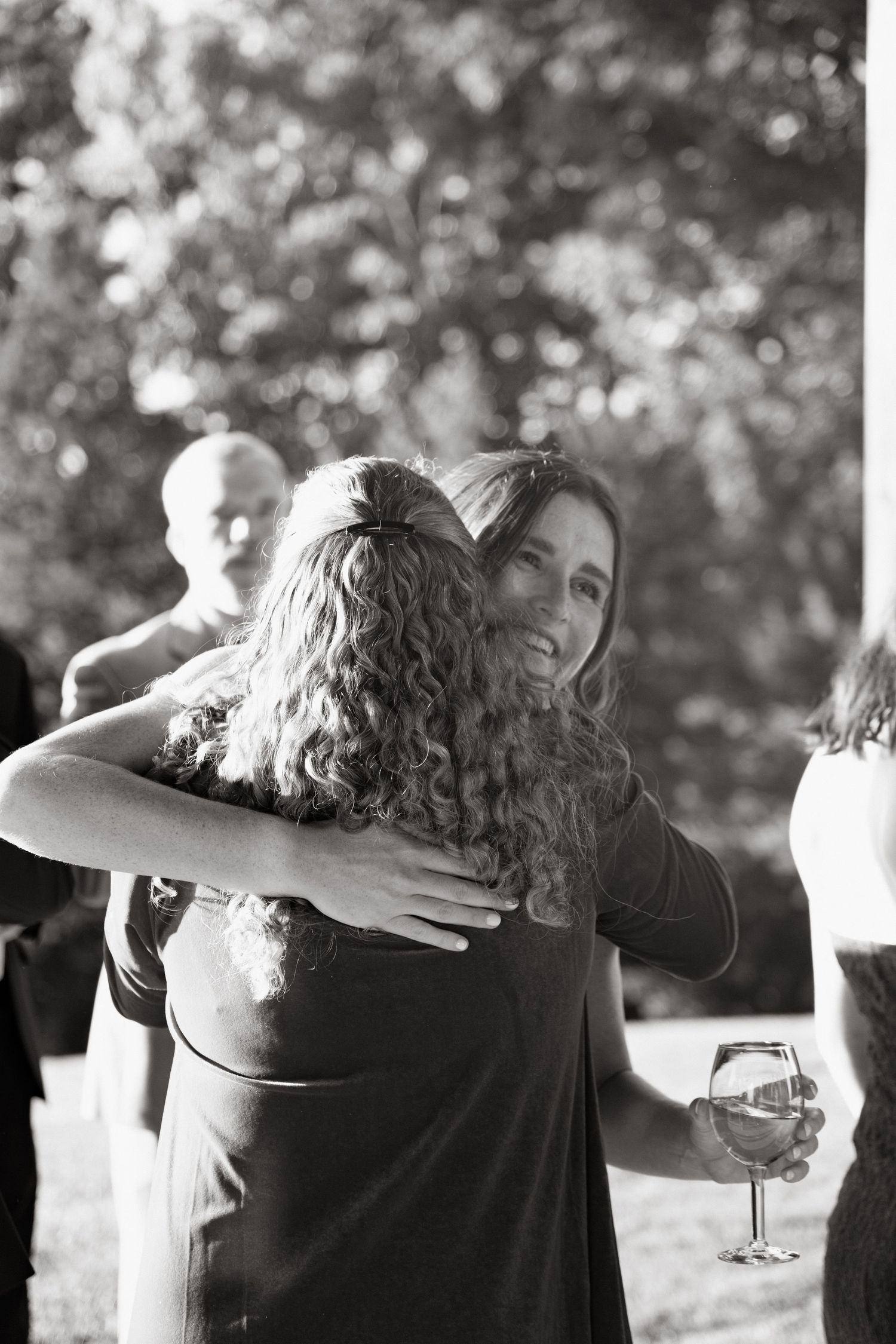 A candid black and white moment of two people embracing at an outdoor gathering while holding wine glasses.