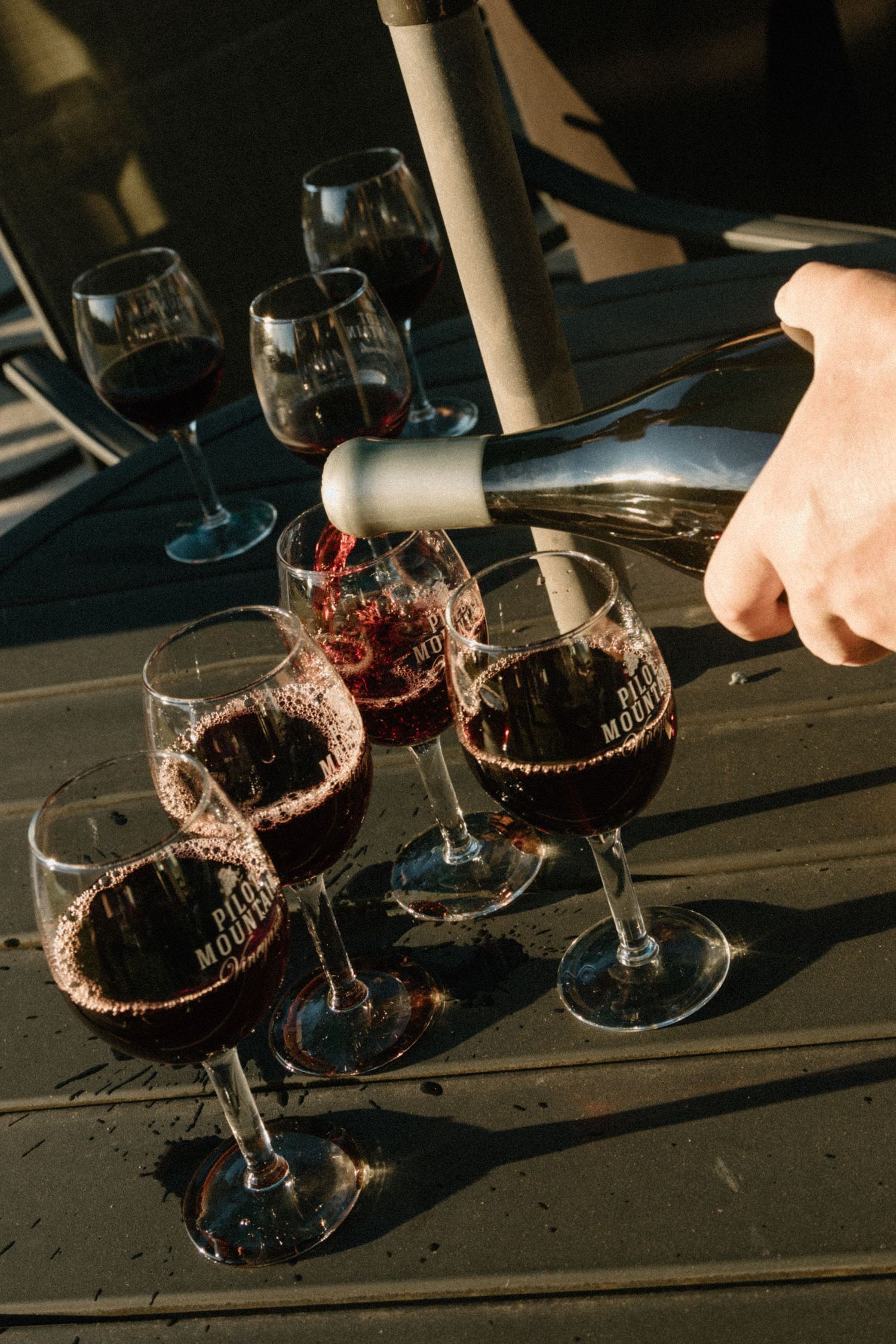 Someone pours red wine into elegant stemmed glasses during a wine tasting on a dark wooden table.