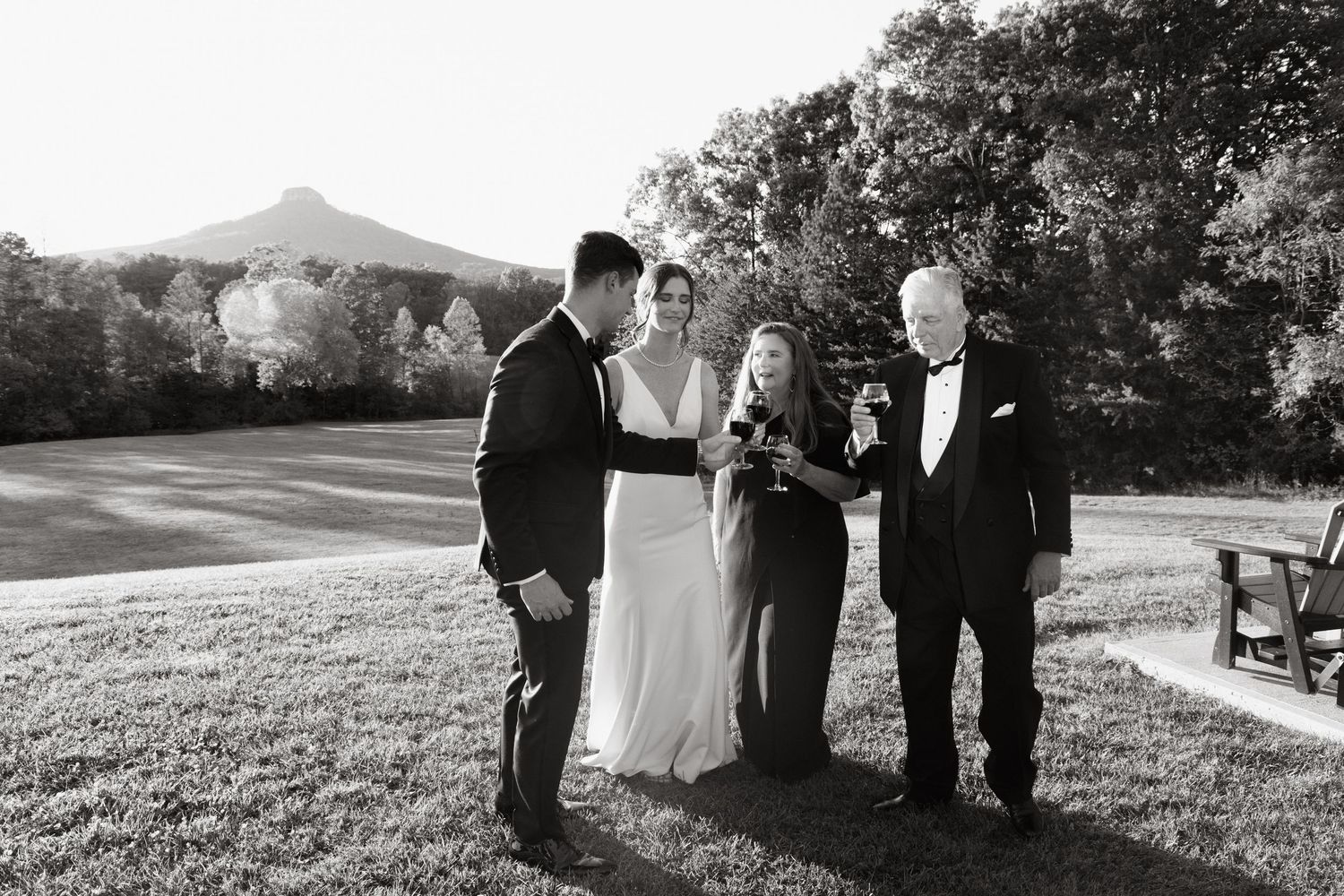 An intimate outdoor wedding ceremony takes place on a grassy hill with trees and buildings in the background.