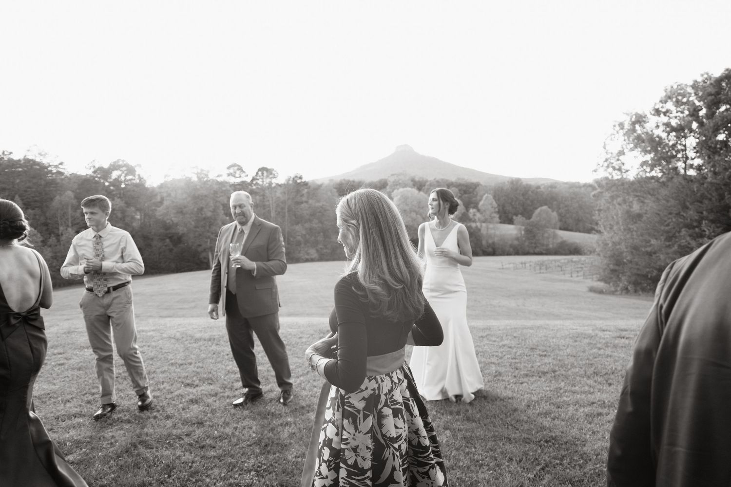 Black and white photo of guests mingling at an outdoor event with mountains visible in the background.