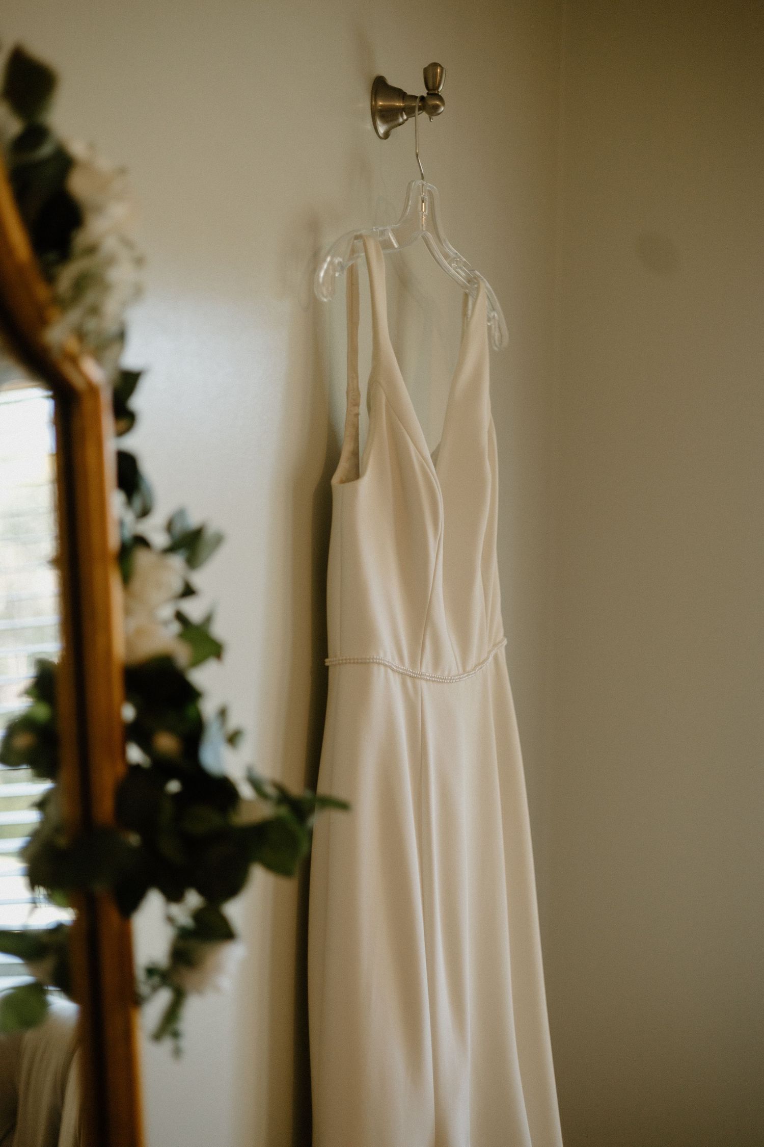 An elegant white wedding dress with spaghetti straps hangs against a neutral wall with greenery decor.