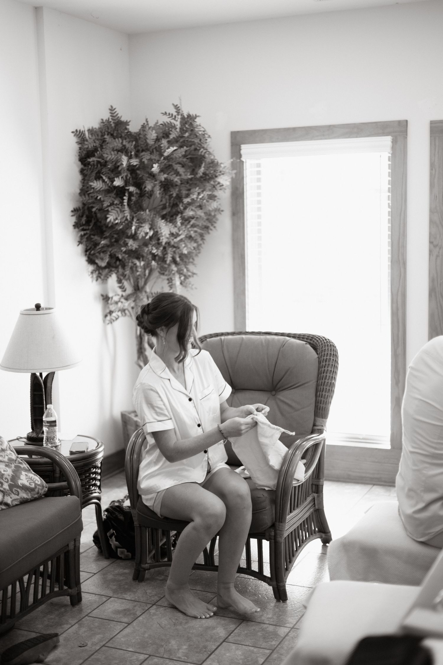 Black and white photo showing someone seated in a wicker chair by a window with a decorative wreath on the wall.