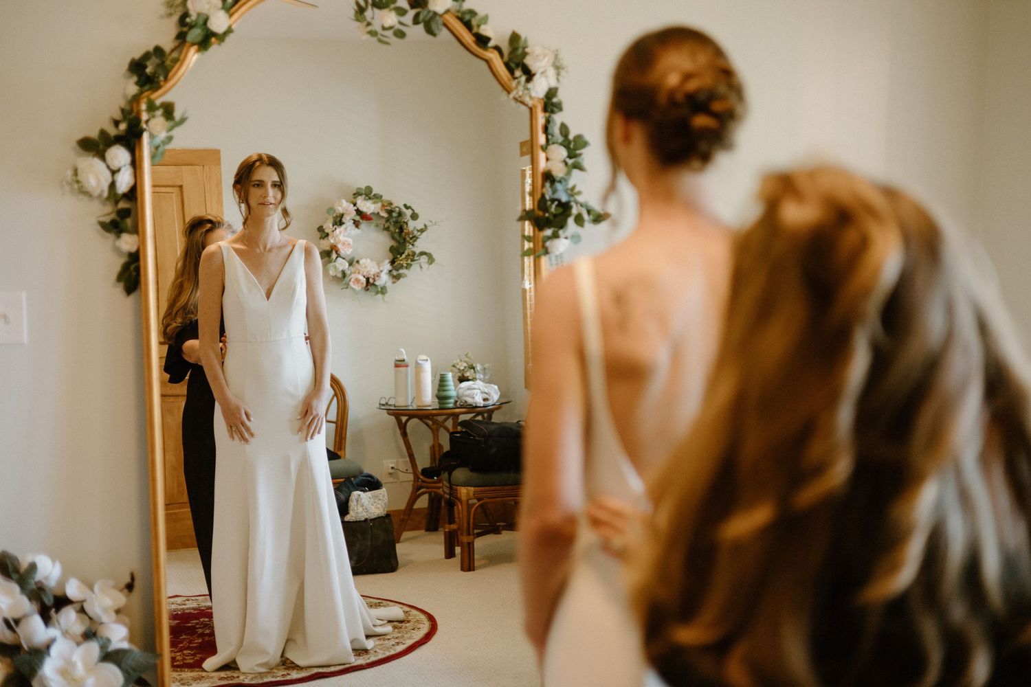 Person in elegant white wedding gown looks at reflection in mirror surrounded by floral arch decorations.