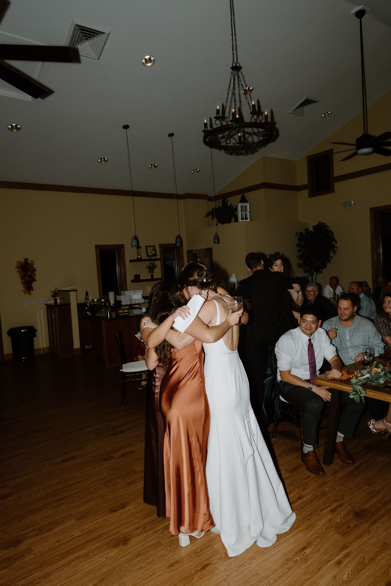 Two people embrace on a wooden dance floor under a chandelier at a reception venue with guests seated nearby.