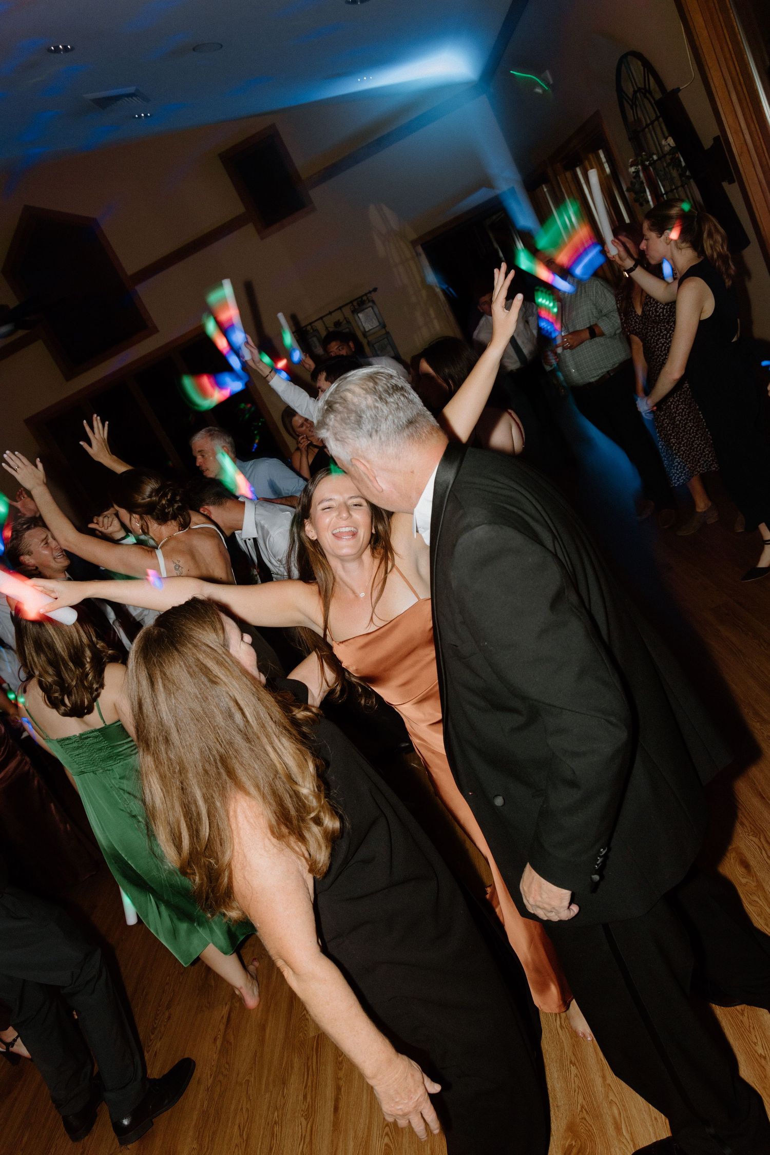 Party guests dancing energetically on a wooden dance floor under colorful disco lights at an indoor event.