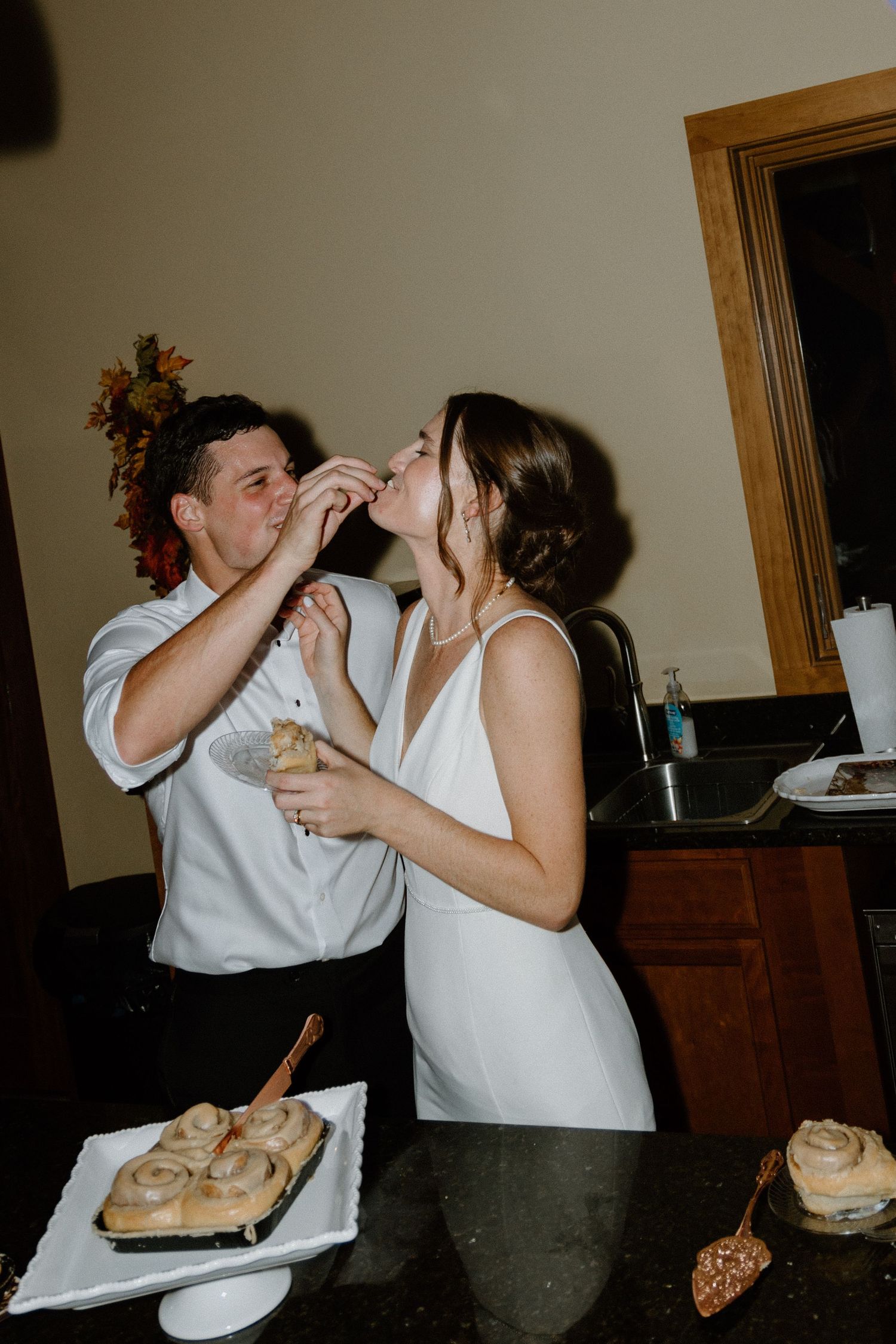 A couple enjoys a playful moment while feeding each other dessert at what appears to be a wedding reception.