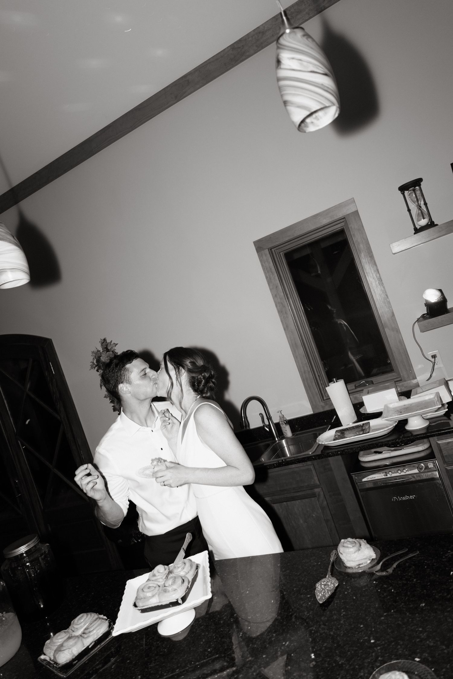 A black and white candid shot of two people dancing and embracing in what appears to be a home setting with pendant lights above.