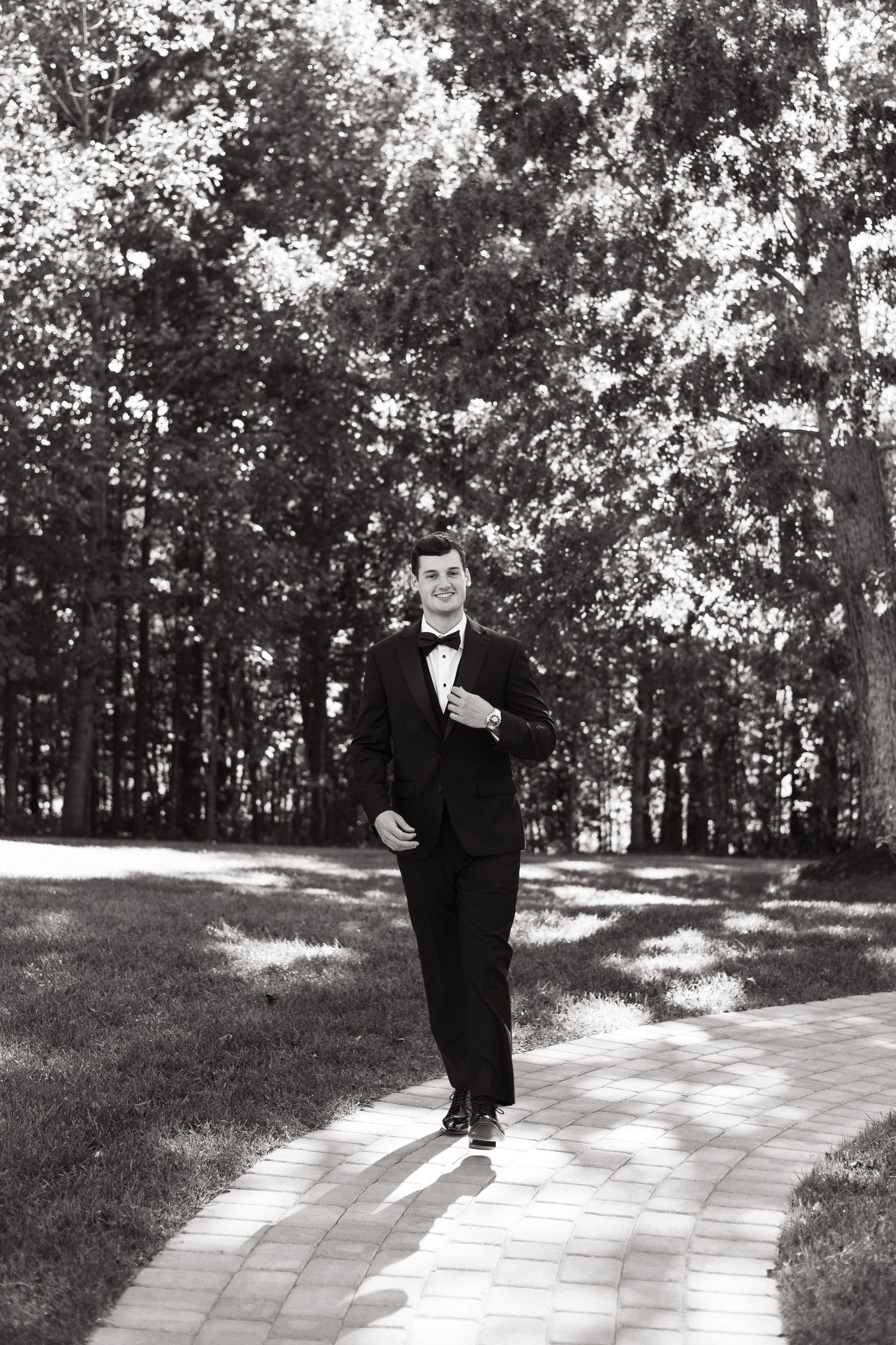 Black and white photo of someone in a formal suit walking along a stone path through a wooded area on a sunny day.