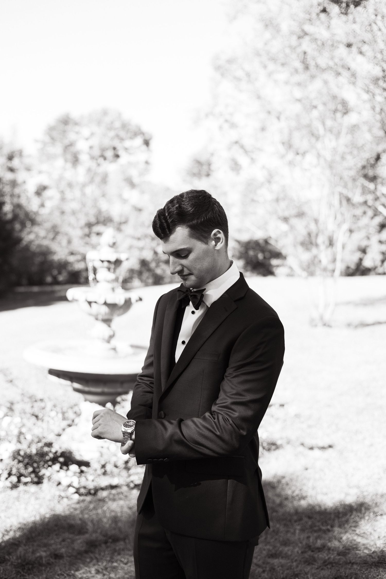 A groom in a black tuxedo looks contemplatively down while standing outdoors in a garden setting.