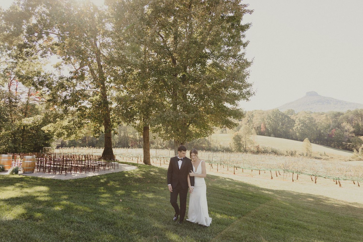 A wedding couple stands together on a grassy hilltop with mountain views and rows of wooden chairs for the ceremony.