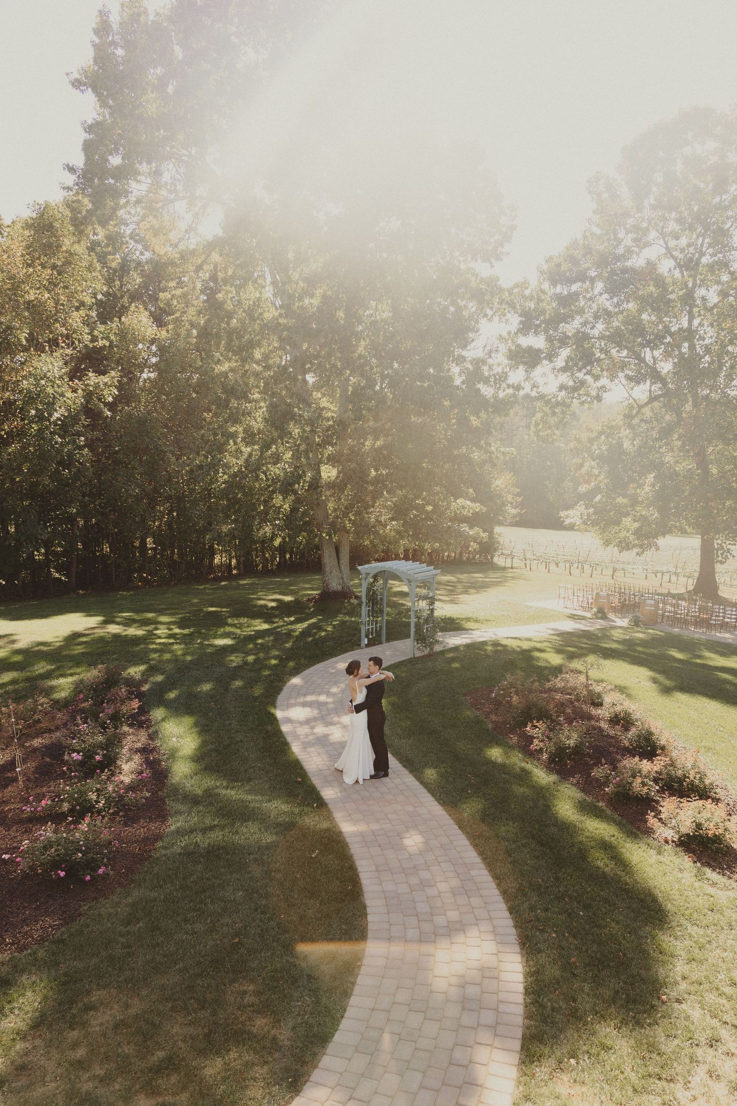 A winding brick pathway through a sunlit garden leads to an elegant outdoor wedding ceremony venue on a misty afternoon.