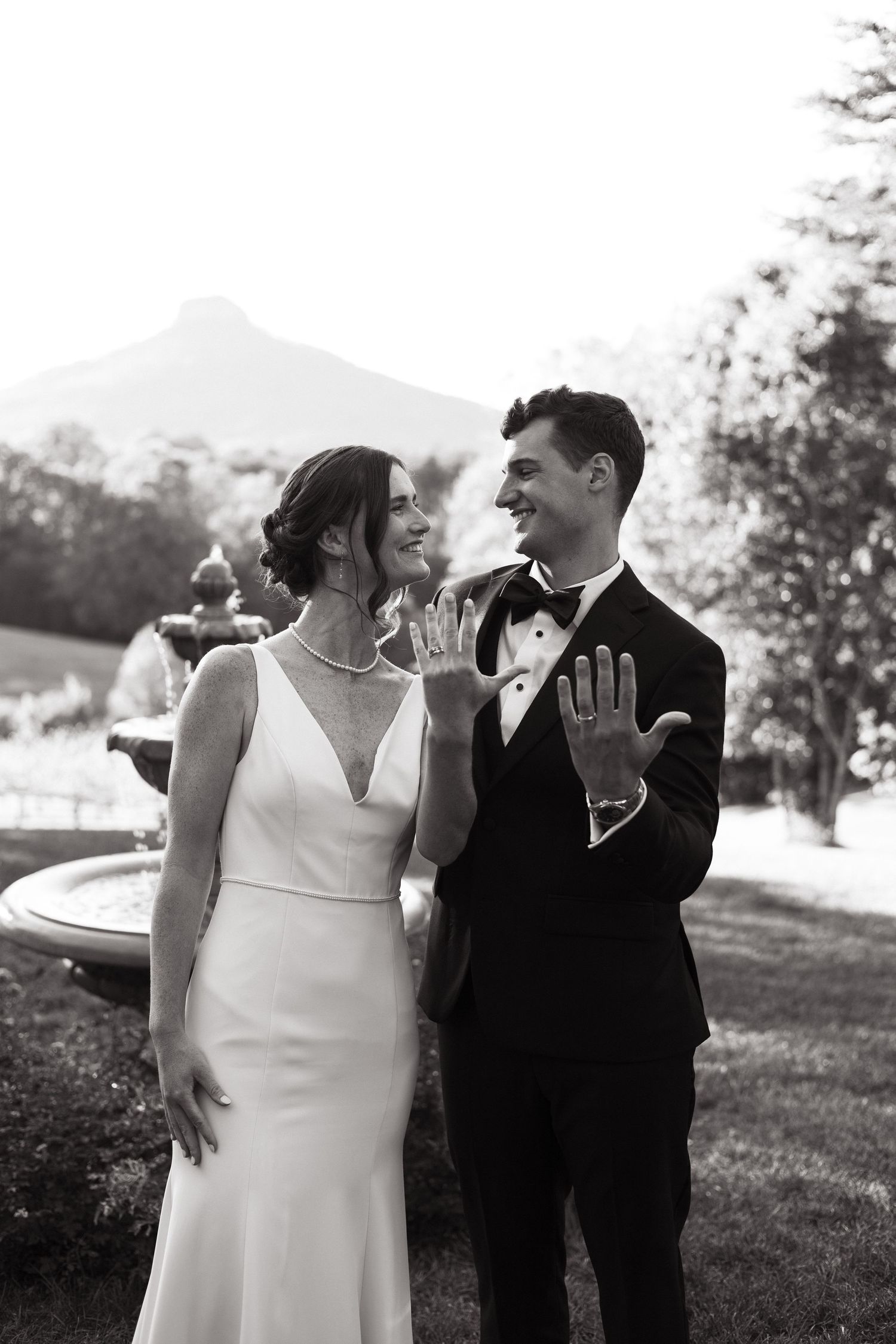 A bride in a fitted white dress and groom in black tuxedo share a candid laugh together outdoors on their wedding day.