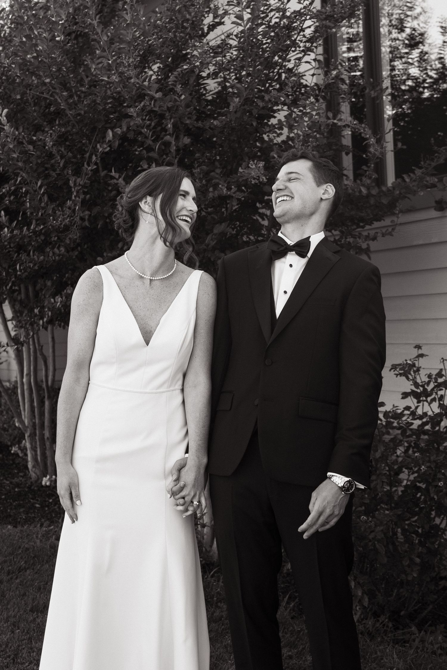 A black and white wedding photo showing a couple in formal attire laughing together outdoors near trees and a building.