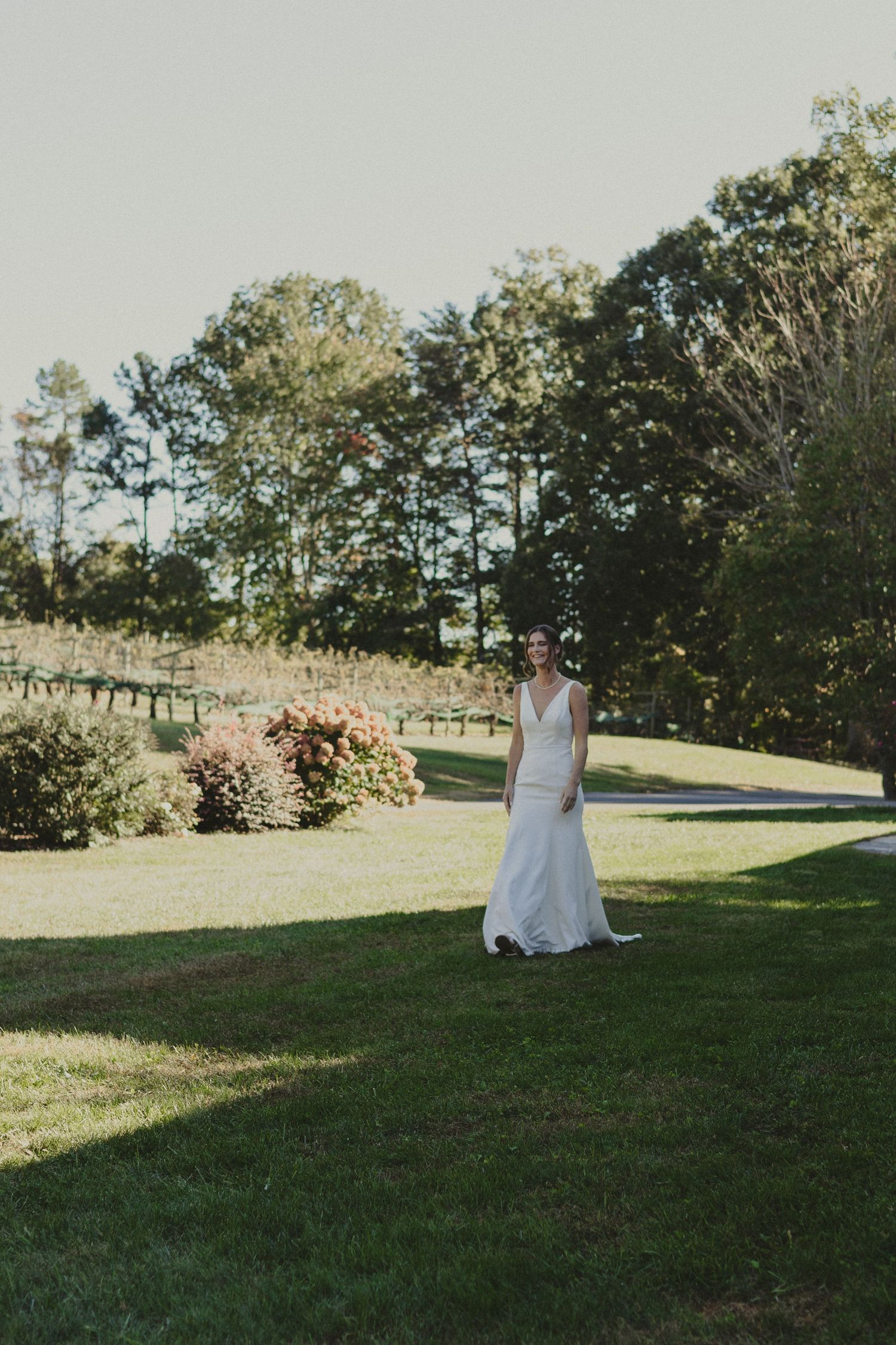 A woman in a white wedding dress walks across a sunlit garden lawn with tall trees in the background.