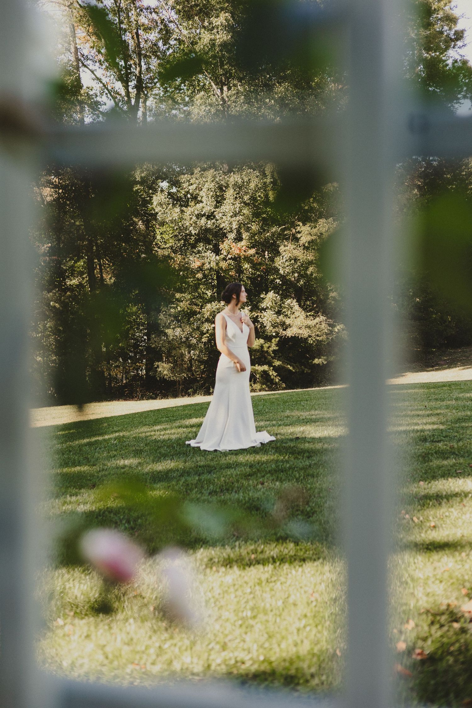 A bride in a white wedding dress stands in a sunlit garden viewed through what appears to be a window or fence.