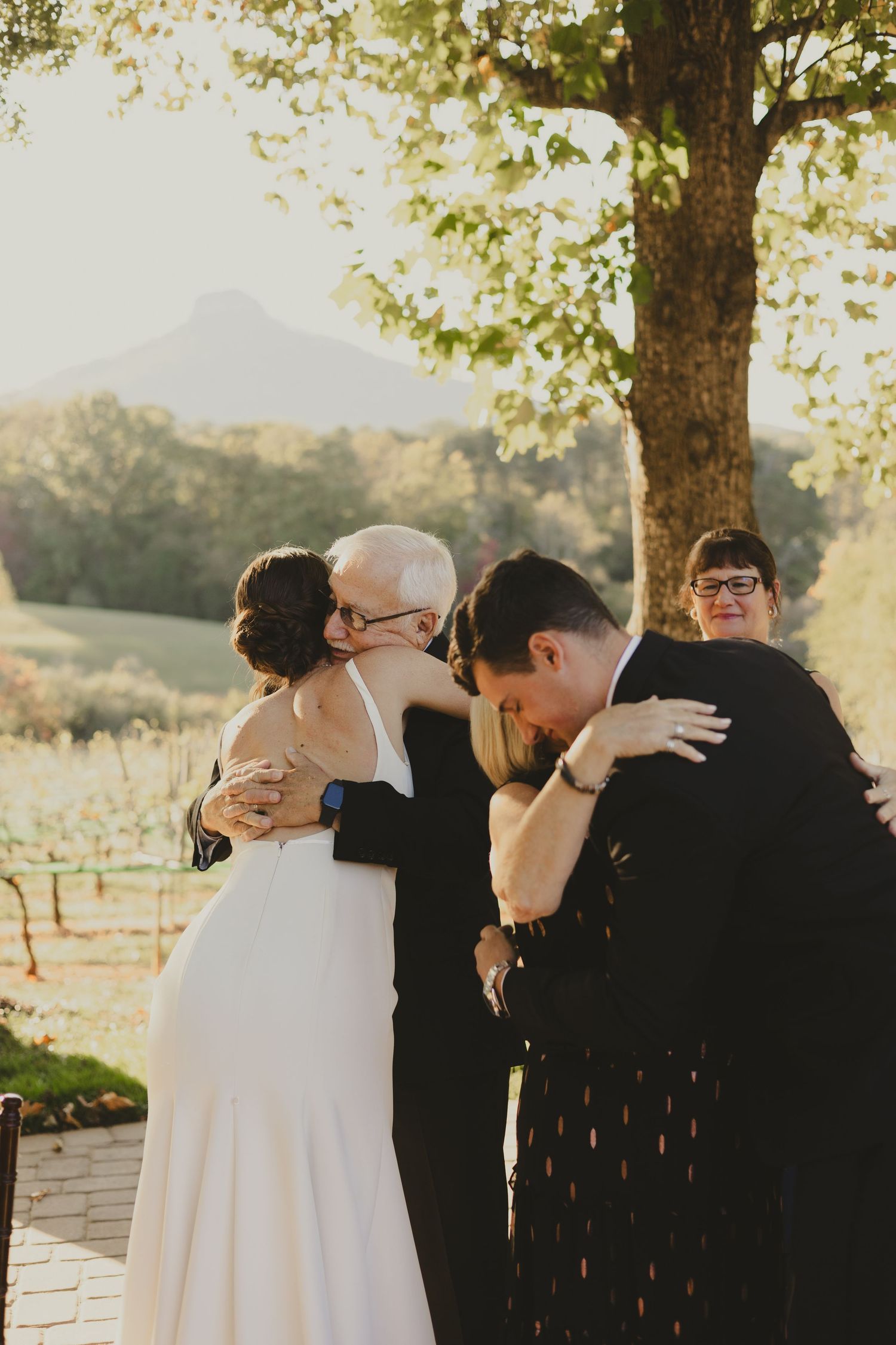 An emotional wedding scene shows a tender family embrace outdoors during sunset at a mountainside venue.
