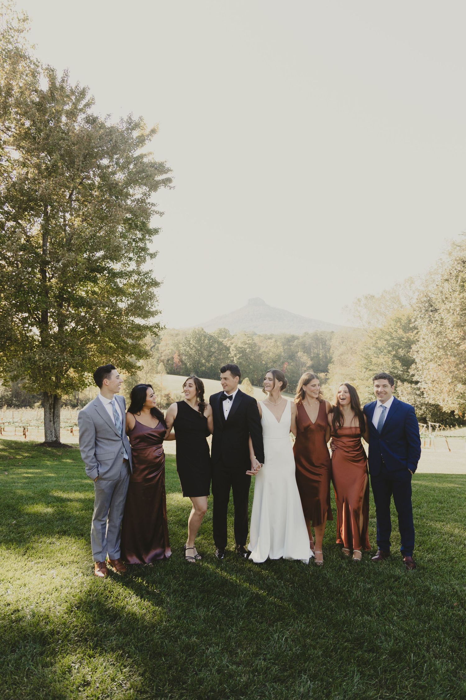Wedding party in formal attire stands together on grassy lawn during autumn outdoor photo session.