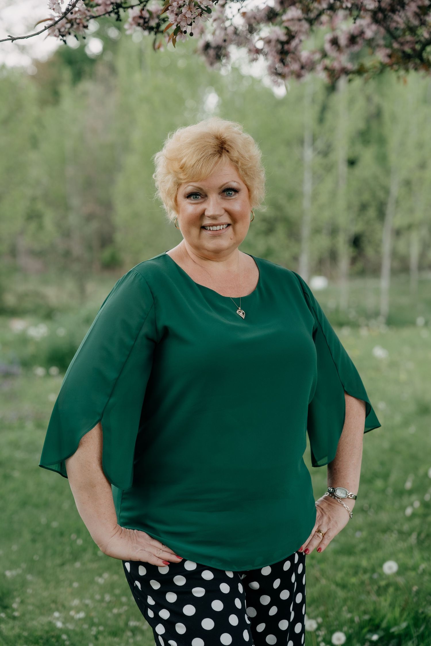 Someone in a green top and polka dot pants poses outdoors near flowering trees in a spring setting.