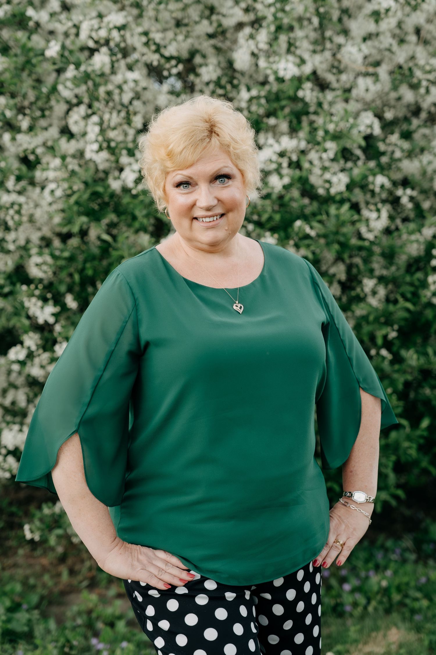 A cheerful woman in a green top and polka dot pants poses outdoors against a blooming white floral backdrop.