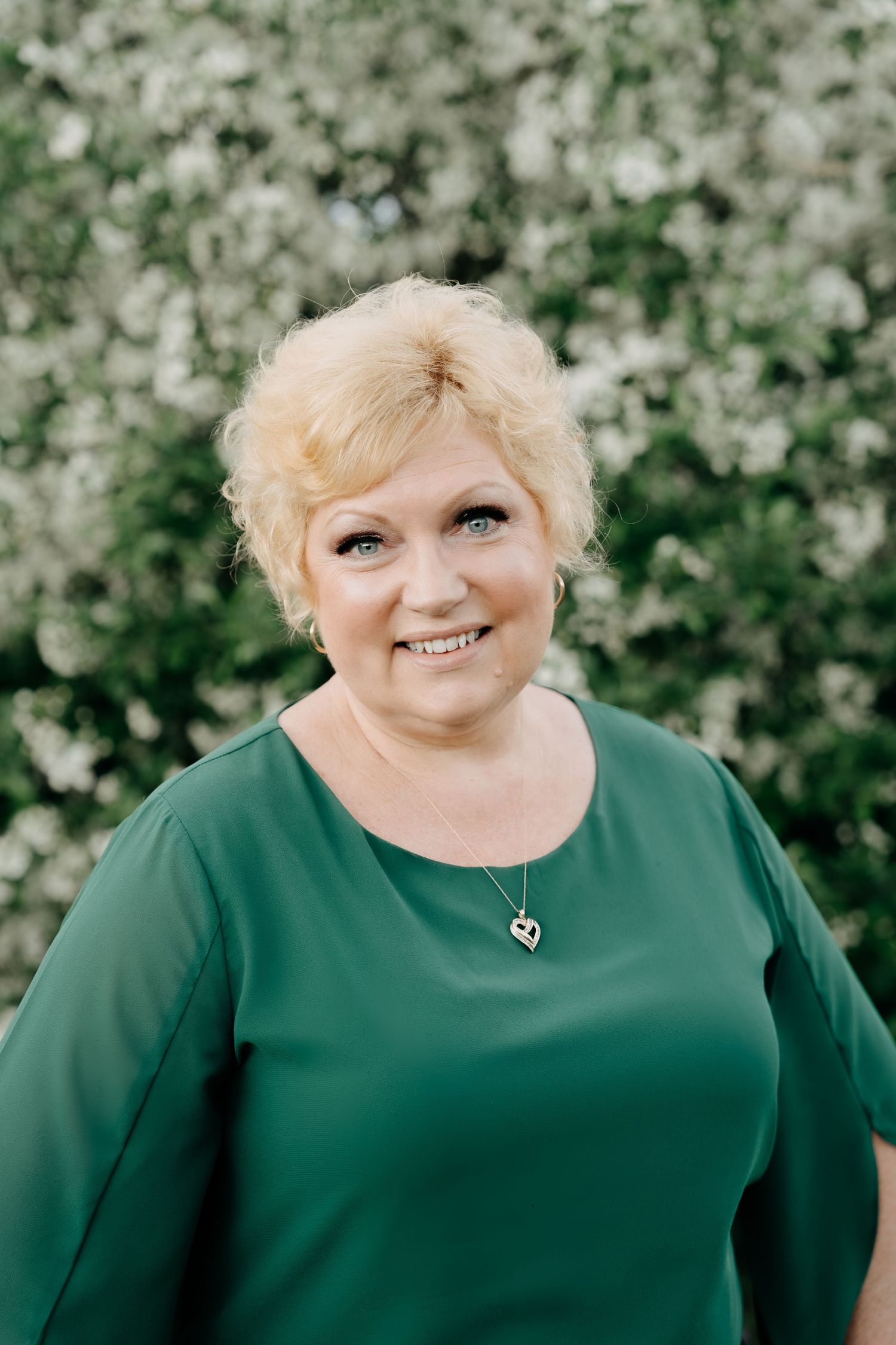A person in an emerald green top poses outdoors against a blooming white flower background wearing a delicate necklace.