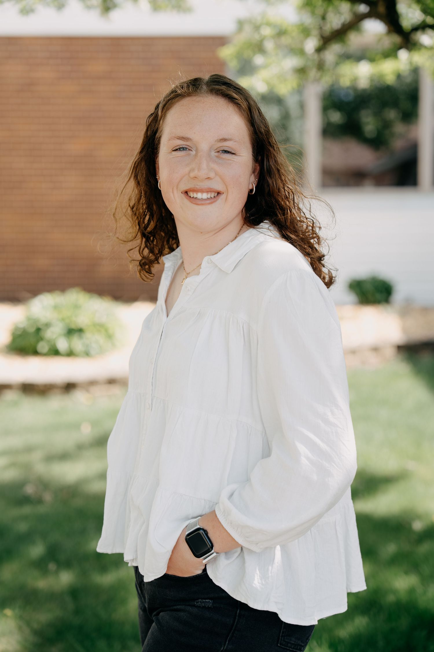 Professional outdoor portrait of someone wearing a white button-down blouse and dark pants on a sunny day.