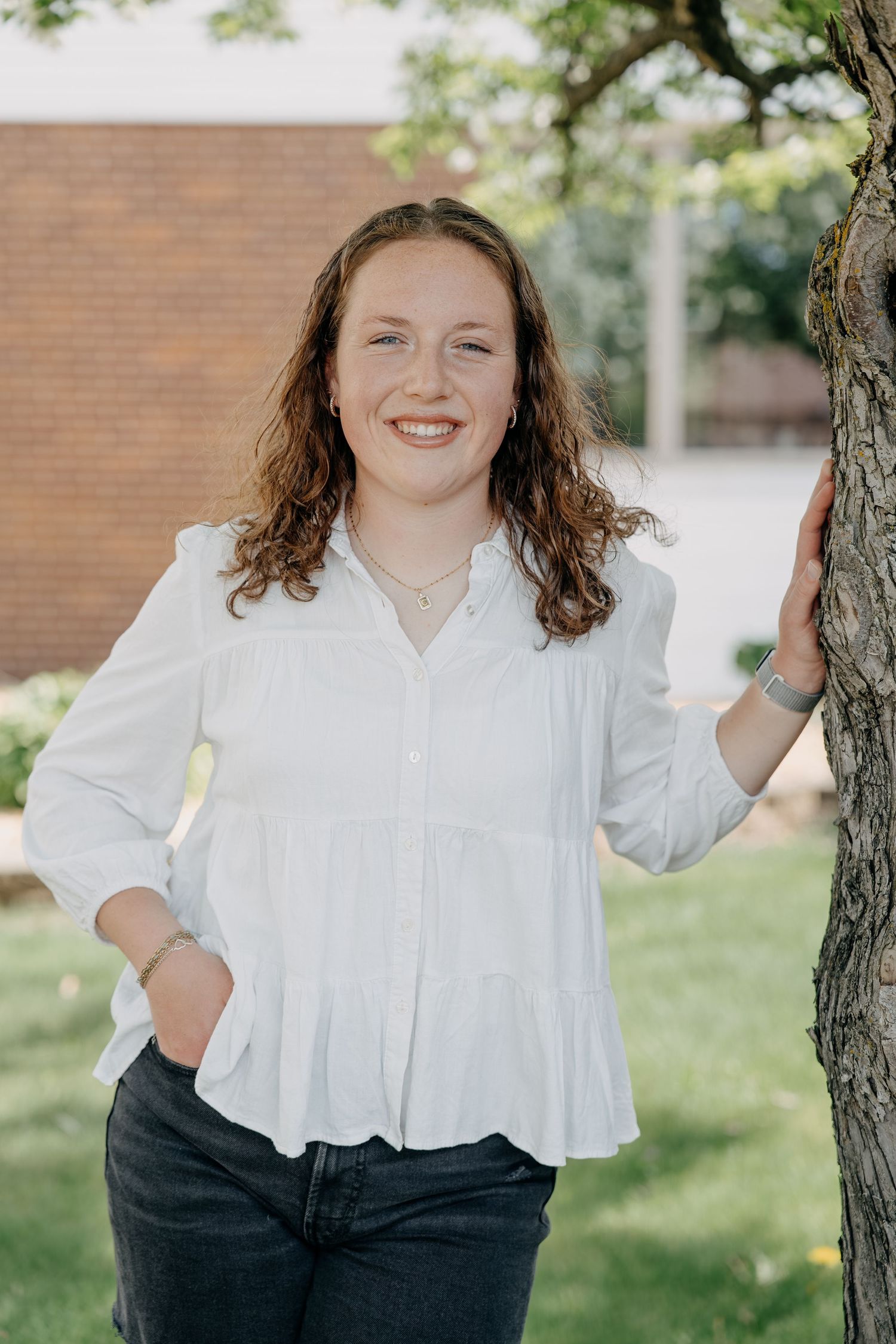 A casual outdoor portrait showing someone in a white button-down blouse and dark jeans standing next to a tree.