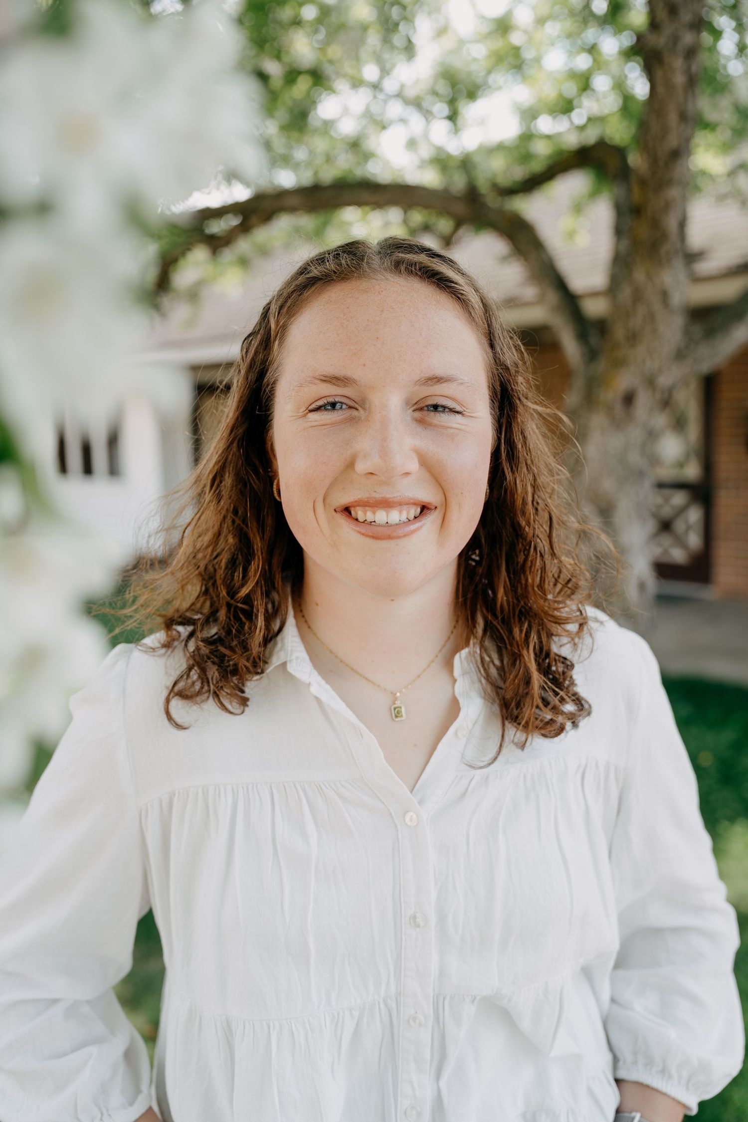 A person wearing a white button-down shirt smiles at the camera while standing outdoors with trees in the background.