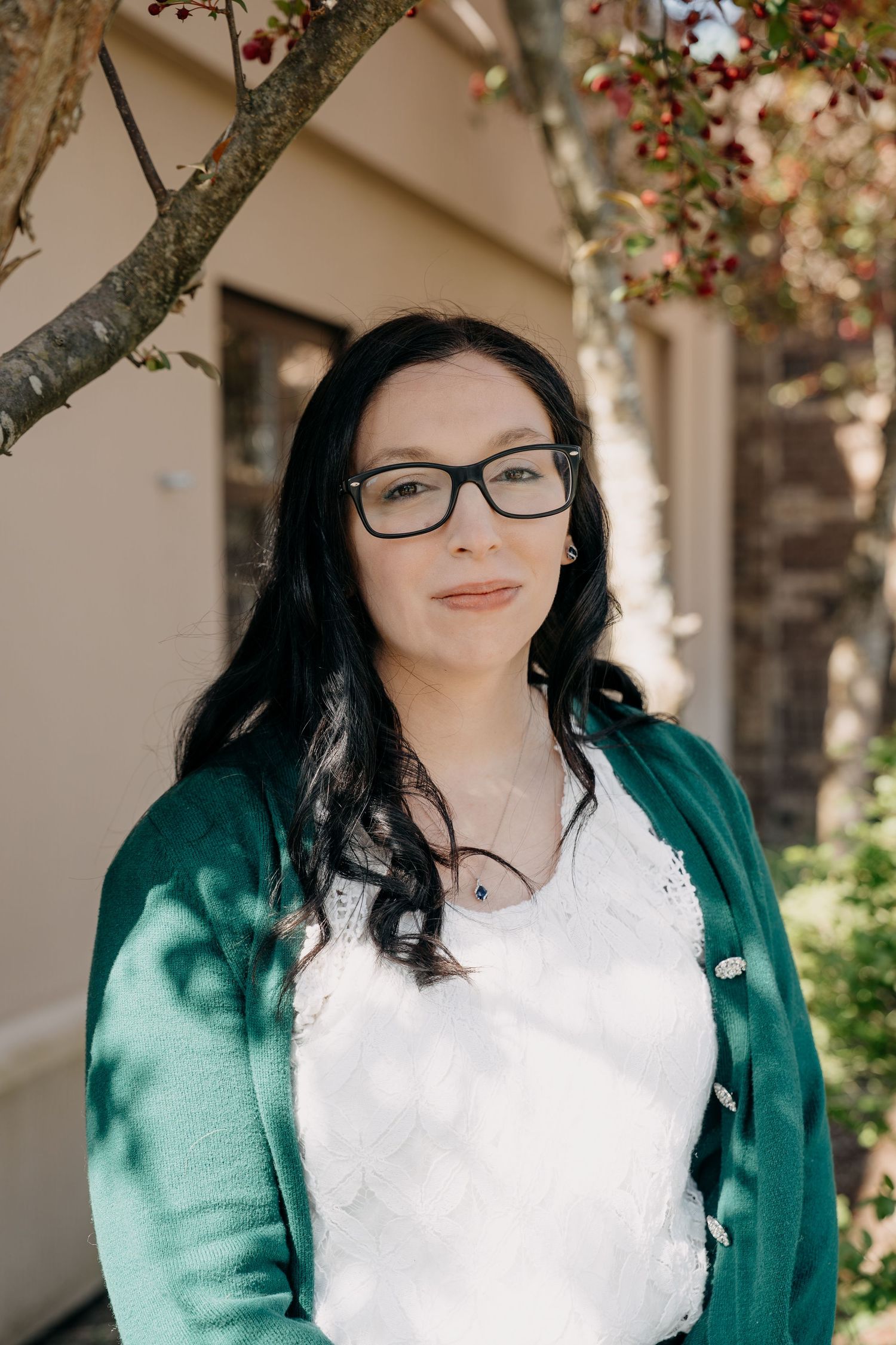 A person with long dark hair and glasses poses outdoors near flowering trees wearing a green cardigan.