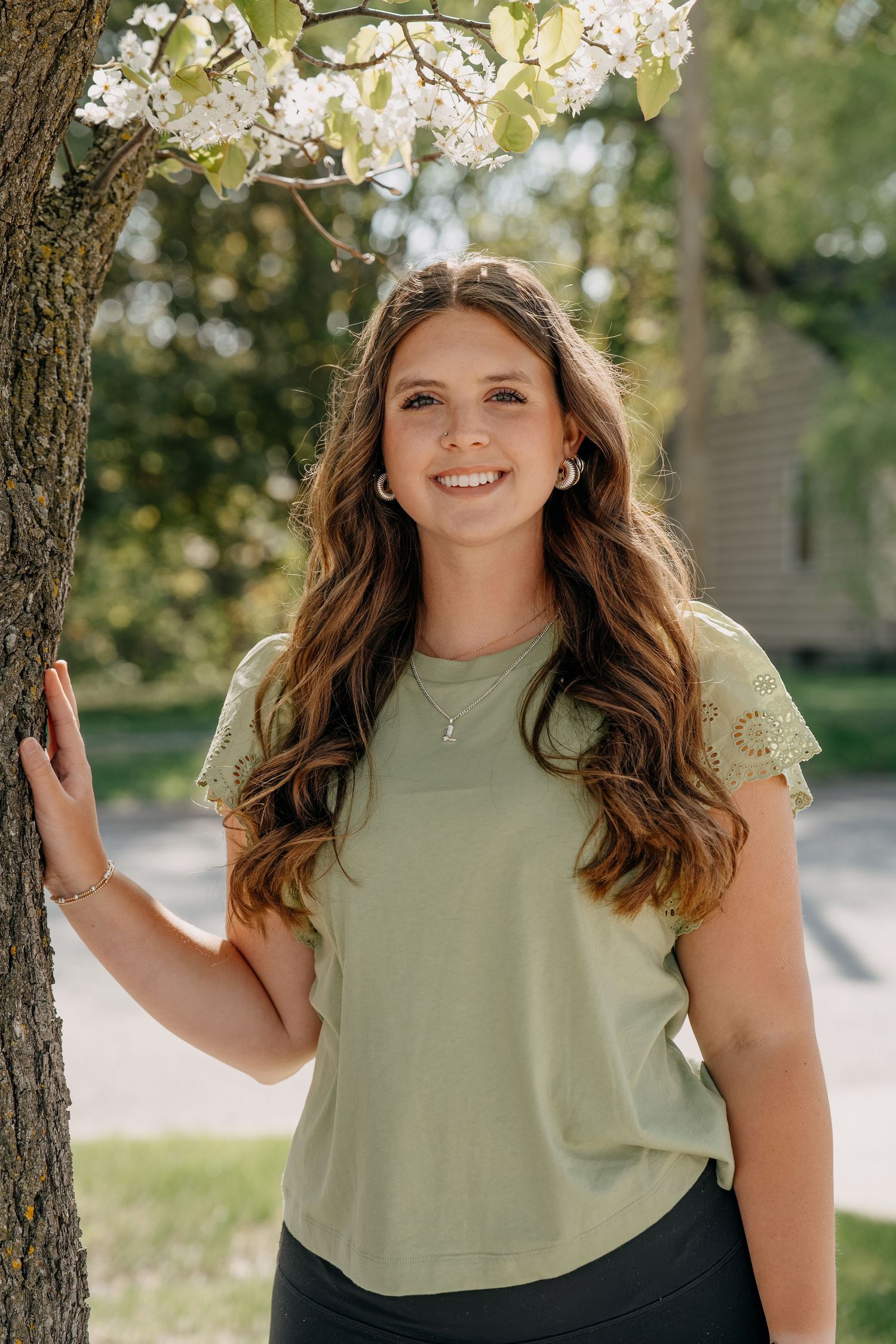 A person in a sage green top stands near a flowering tree on a sunny spring day in an outdoor setting.