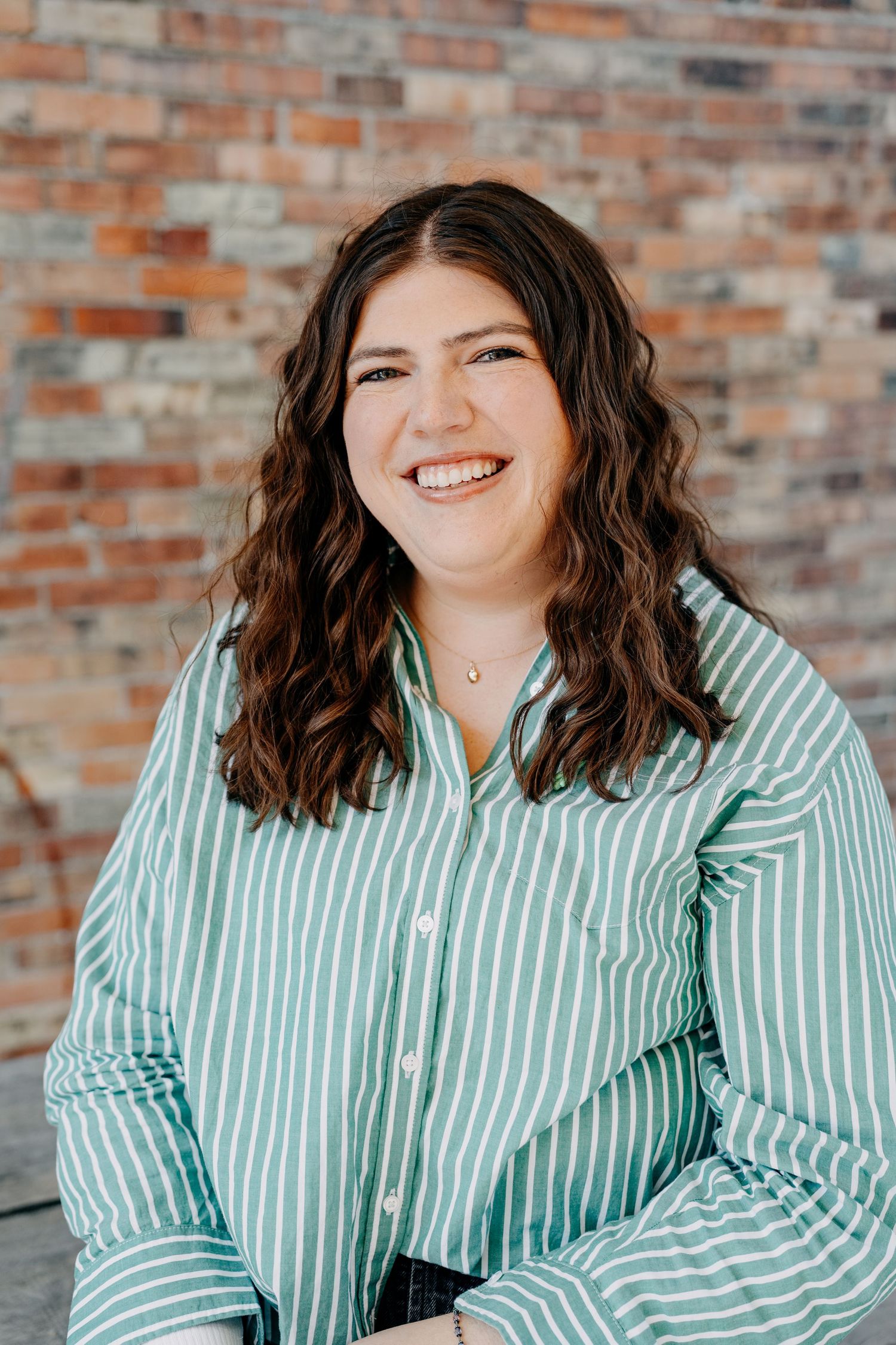 A person with wavy brown hair wearing a mint green and white striped button-down shirt poses against a brick wall.