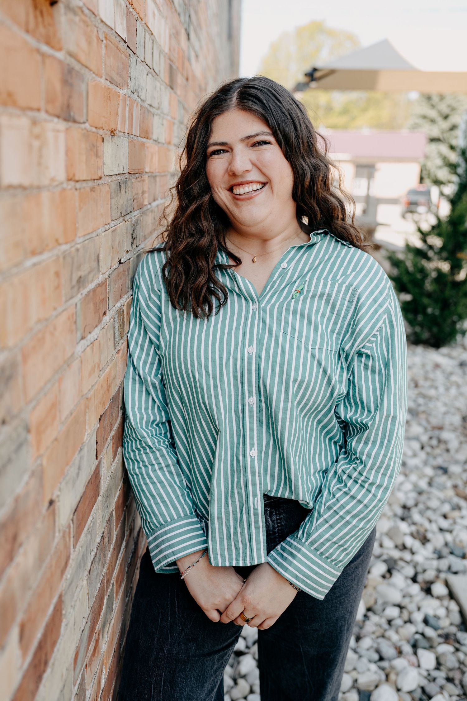 Smiling figure in a mint green striped button-down shirt poses against a rustic brick wall.