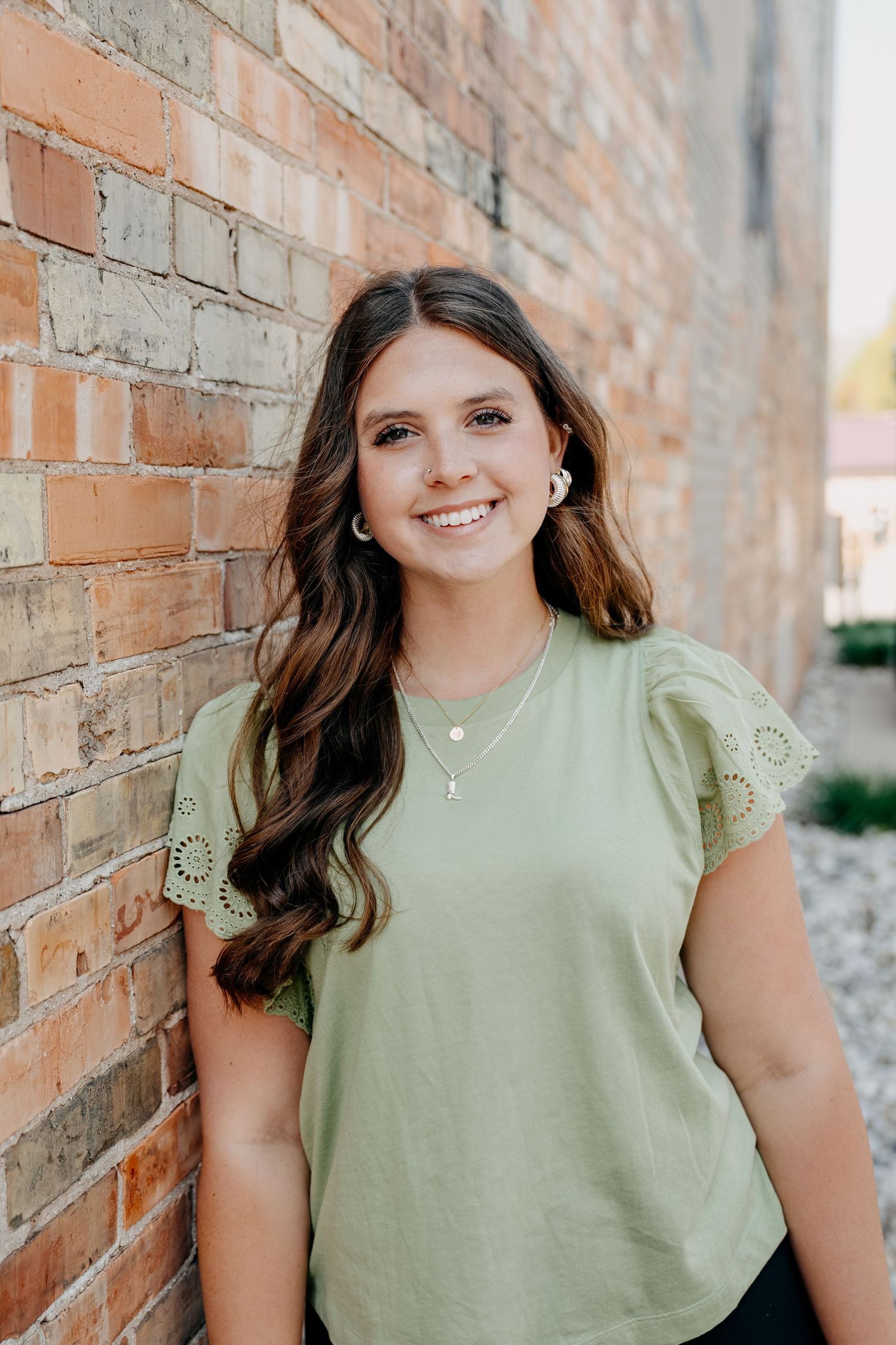 Woman in sage green lace-sleeve top smiles while standing against rustic brick wall outdoors.