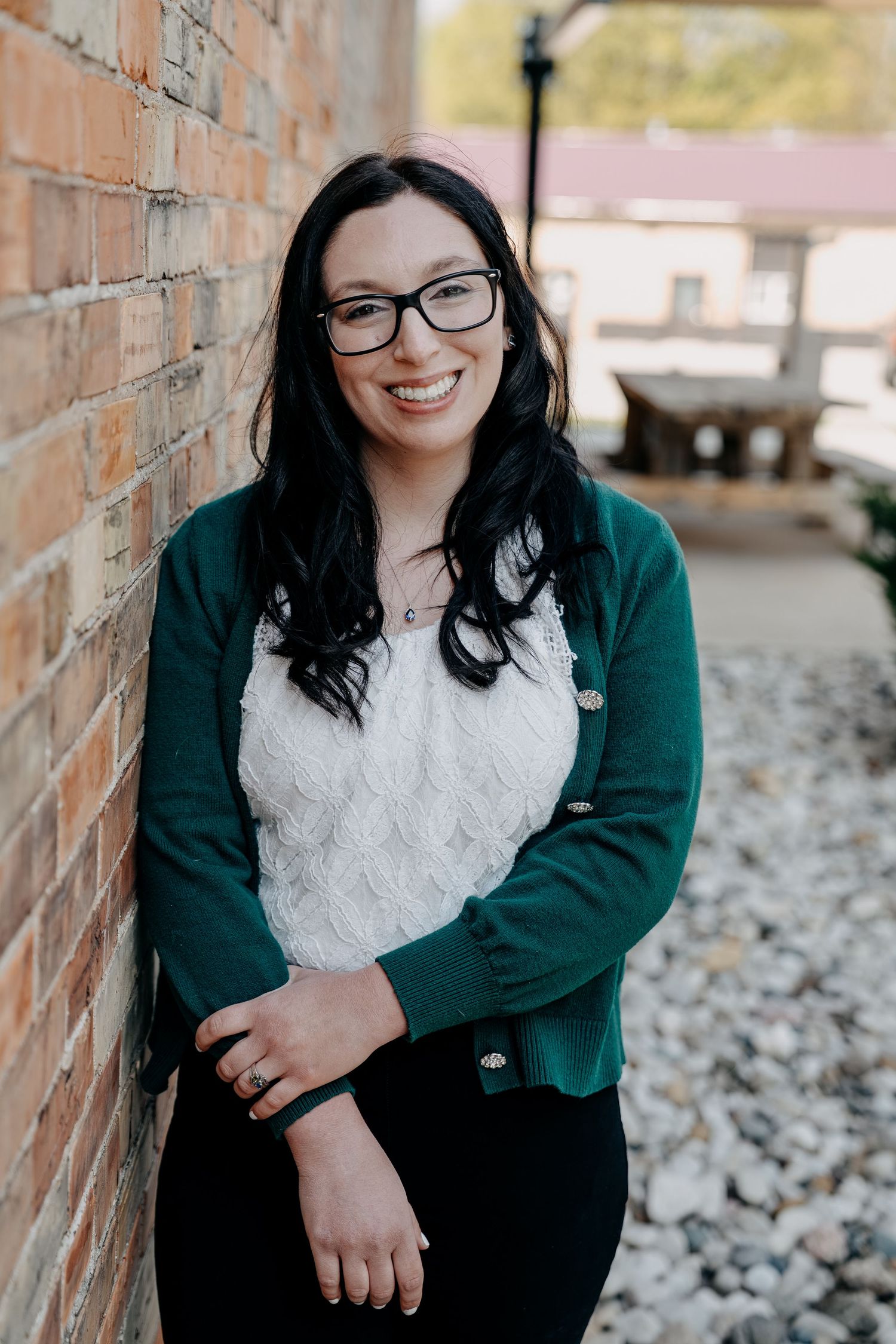 A person in glasses and teal cardigan smiles while leaning against a brick wall outdoors.