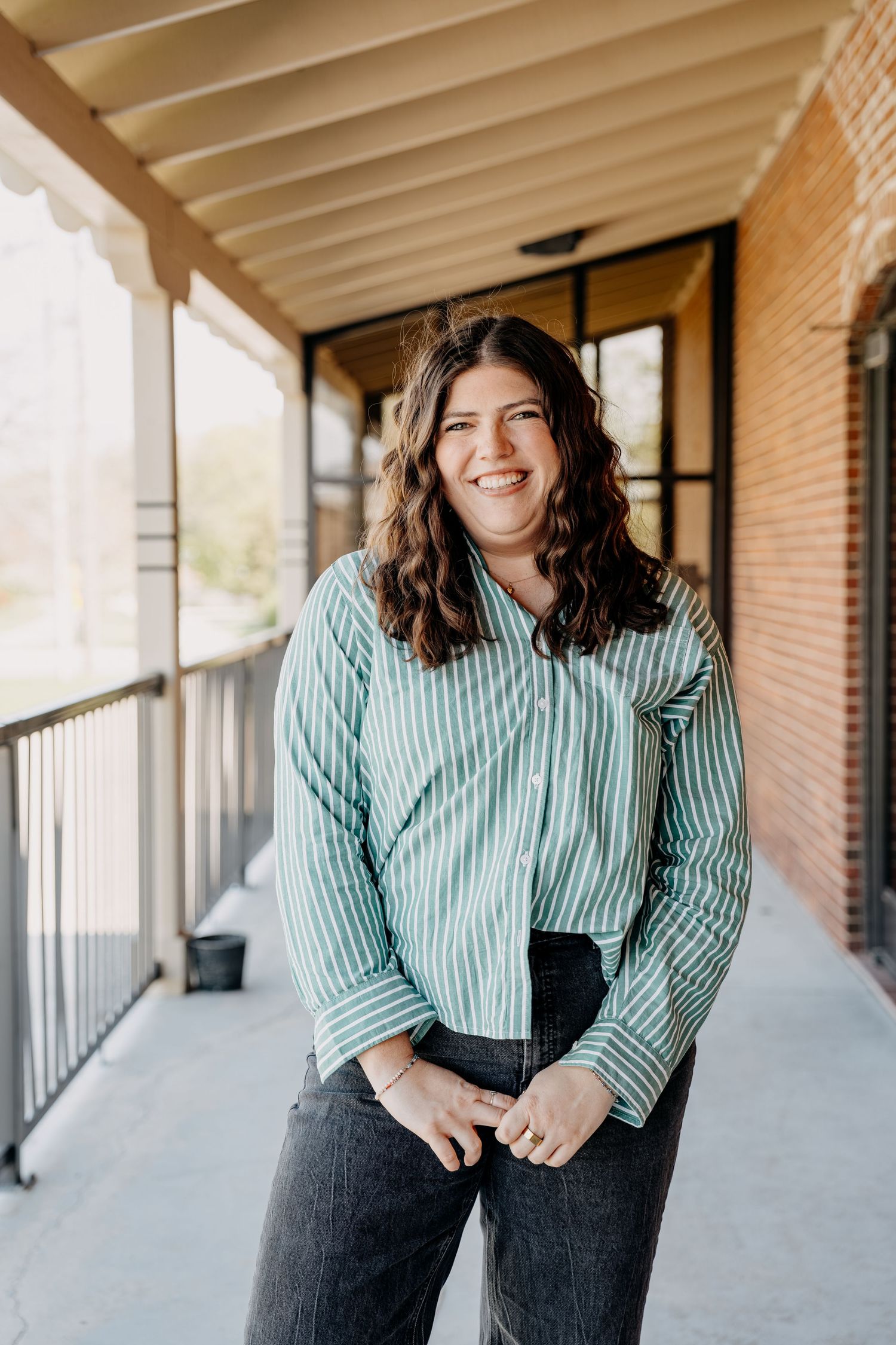 A woman wearing a striped button-up shirt stands smiling on a covered porch with brick columns.