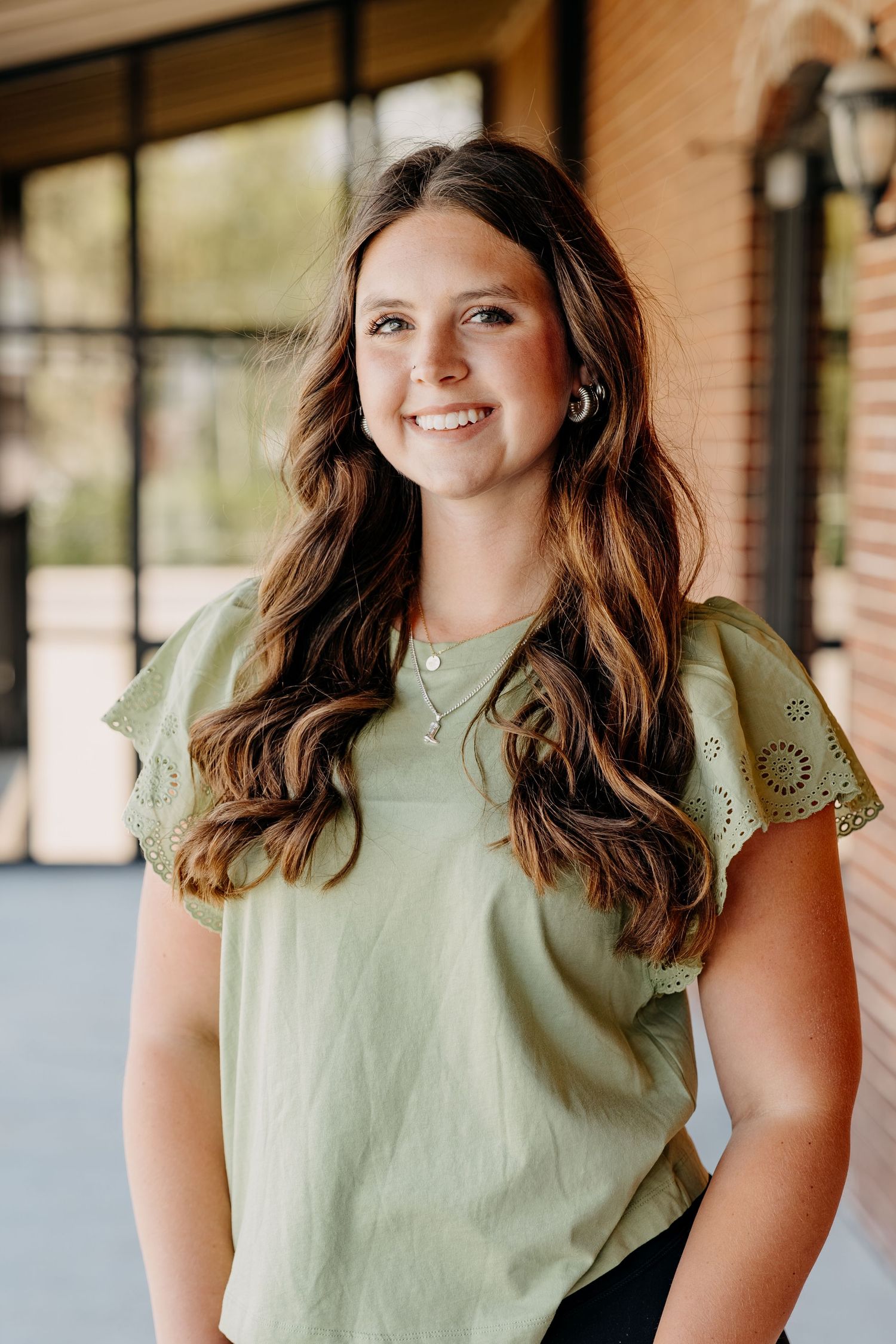 Person with long brown wavy hair wearing a sage green t-shirt poses against brick background.