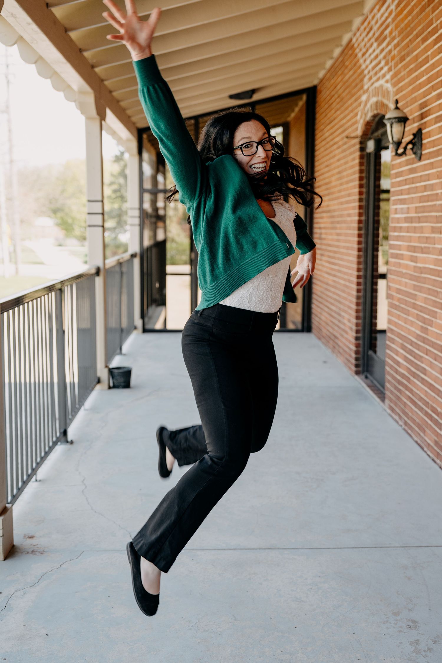 Energetic person in green sweater and black pants jumping joyfully on outdoor walkway with brick wall background.