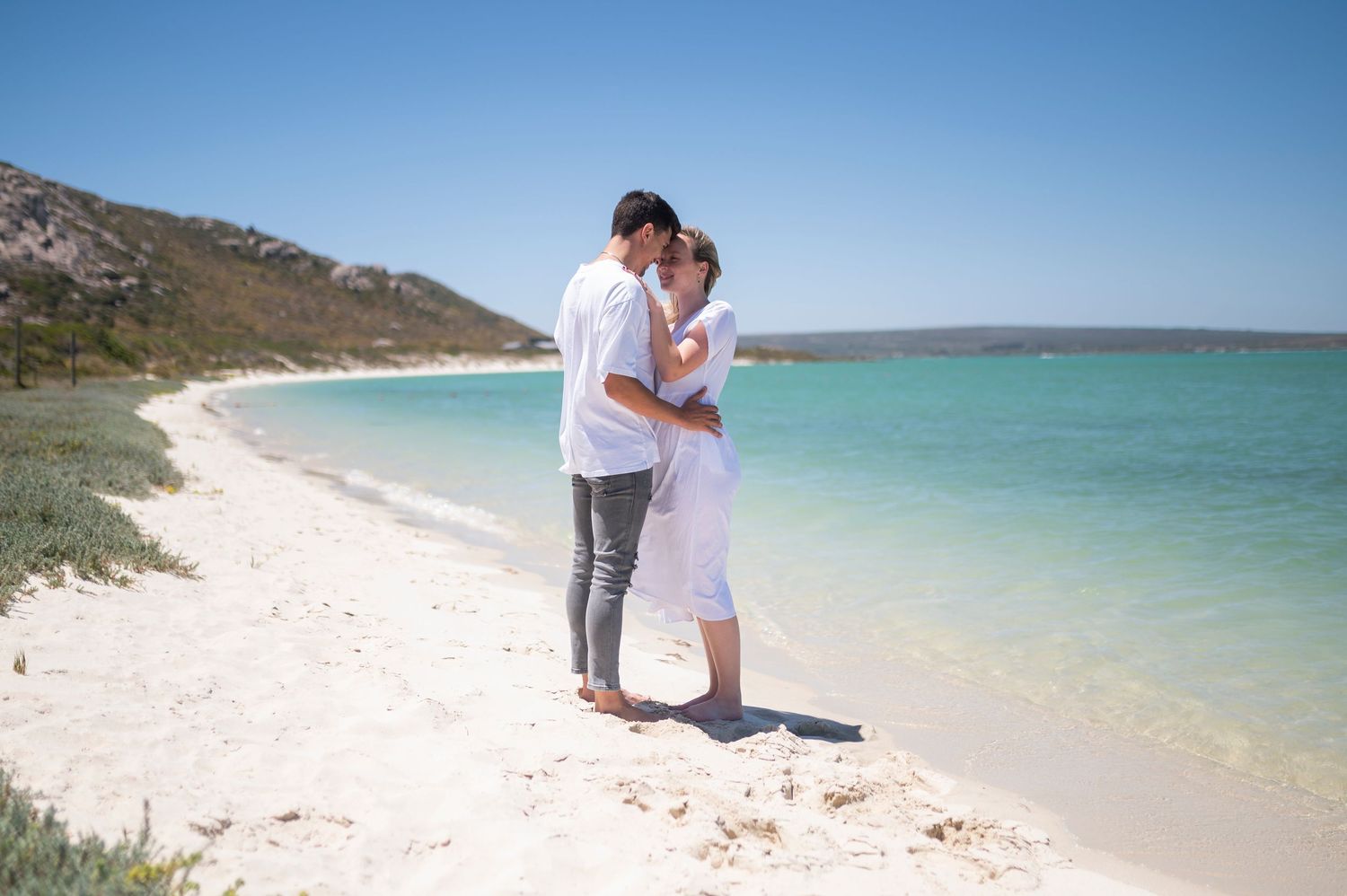 A couple embraces on a pristine white sand beach with turquoise waters in the background.