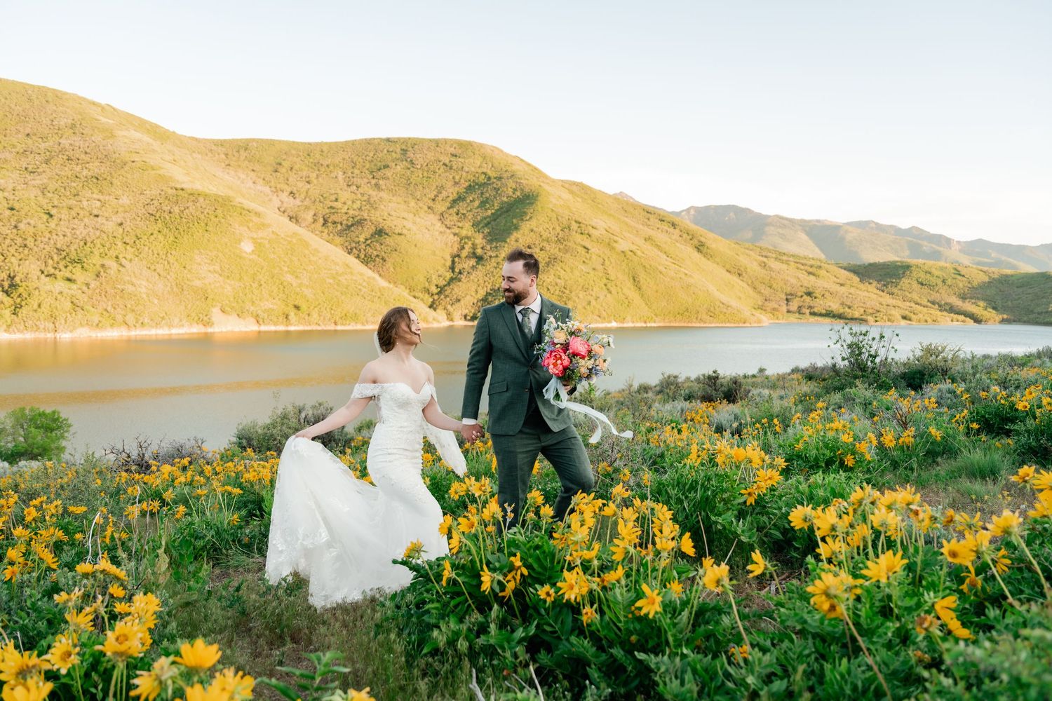 Couple walks through vibrant yellow wildflowers overlooking scenic mountain lake during golden hour.
