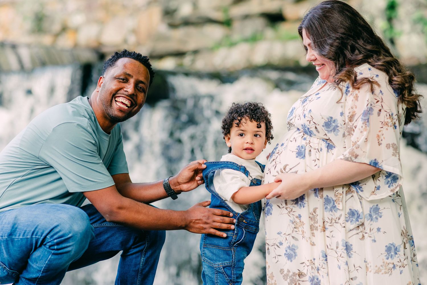 Expectant parents in casual outfits share a joyful moment with their toddler against the waterfall at Southford Falls, CT