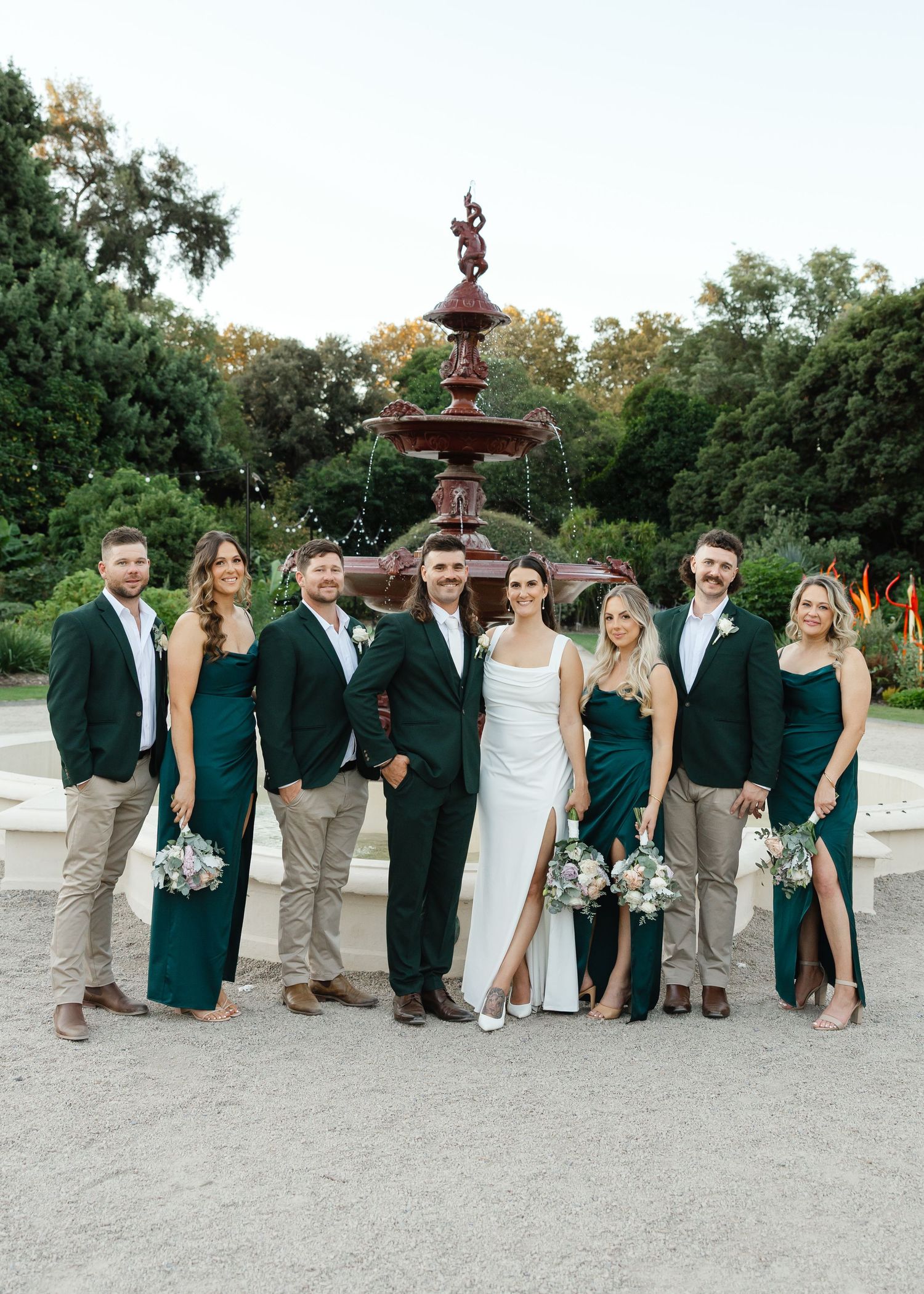 Wedding party in emerald green and khaki attire poses in front of ornate fountain in garden setting.
