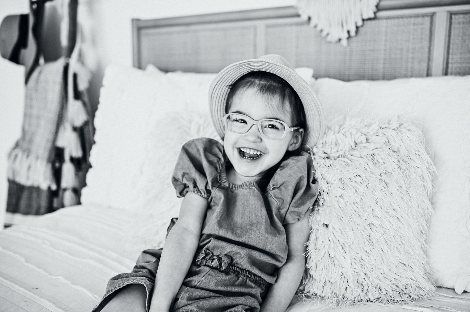 Black and white image of happy young girl wearing round glasses and sun hat on bed.