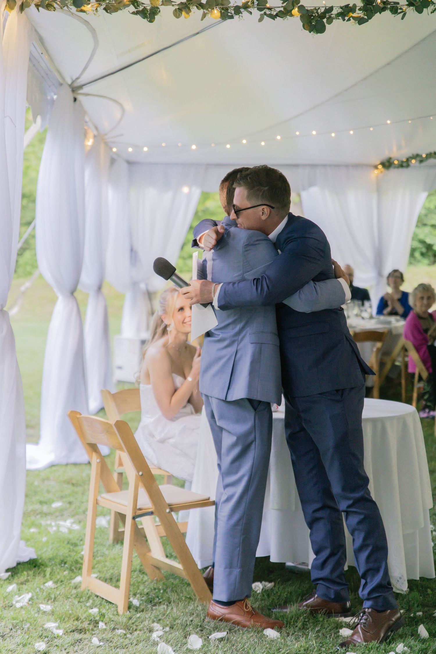 Two people embrace during a wedding reception speech under a decorated white tent with string lights and greenery.