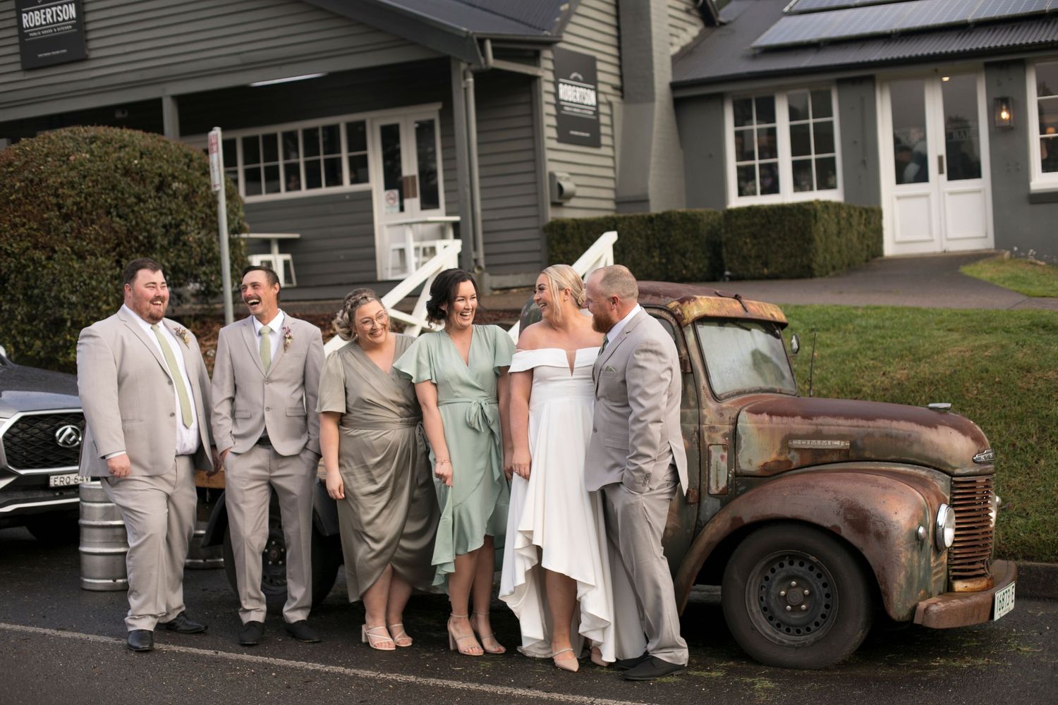 A wedding party poses in front of a rustic vintage truck wearing light colored formal attire creating a timeless scene.