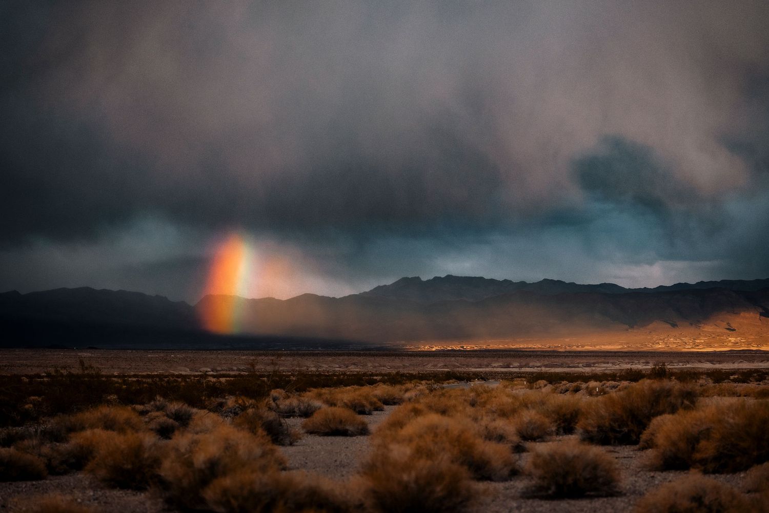 Vivid rainbow emerges from dark storm clouds over arid desert valley landscape.