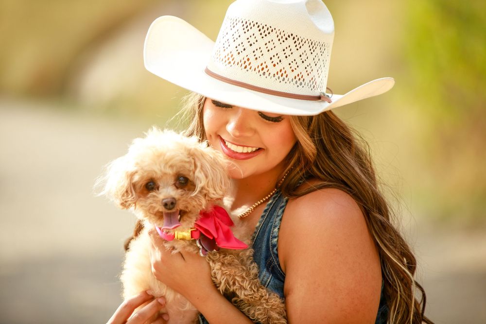Someone in a white cowboy hat and denim top smiling while holding a small fluffy brown dog with a pink bow.