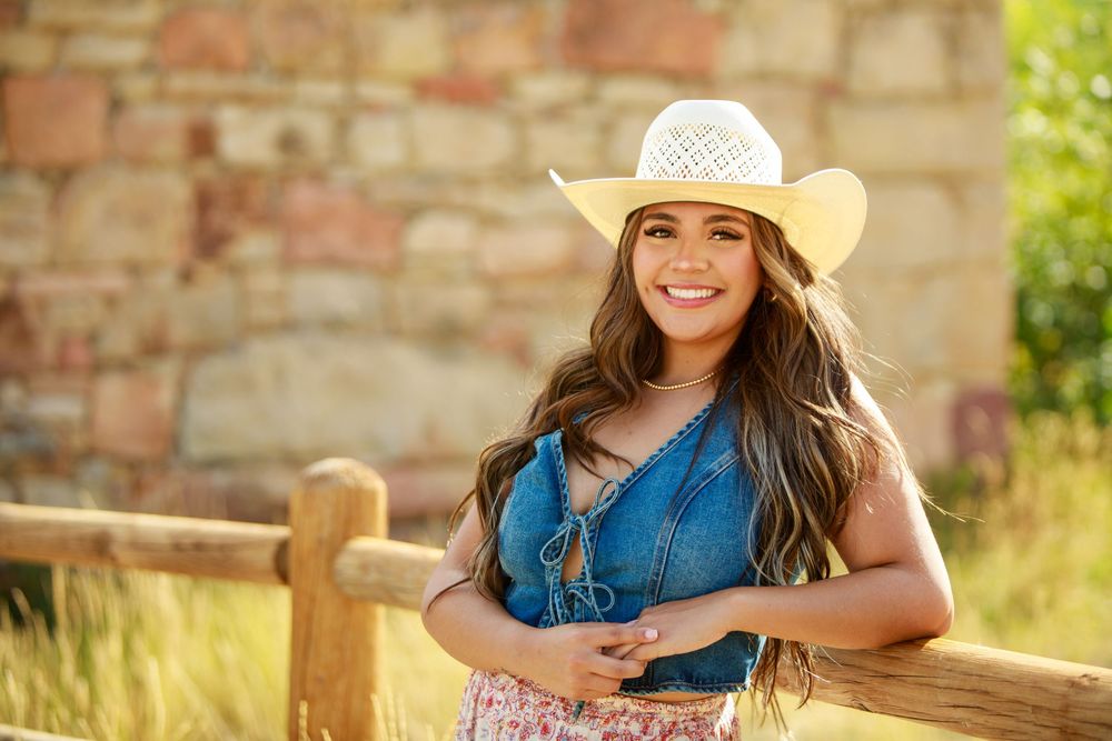 A person wearing a straw cowgirl hat and denim top poses next to a wooden fence post in bright sunlight.