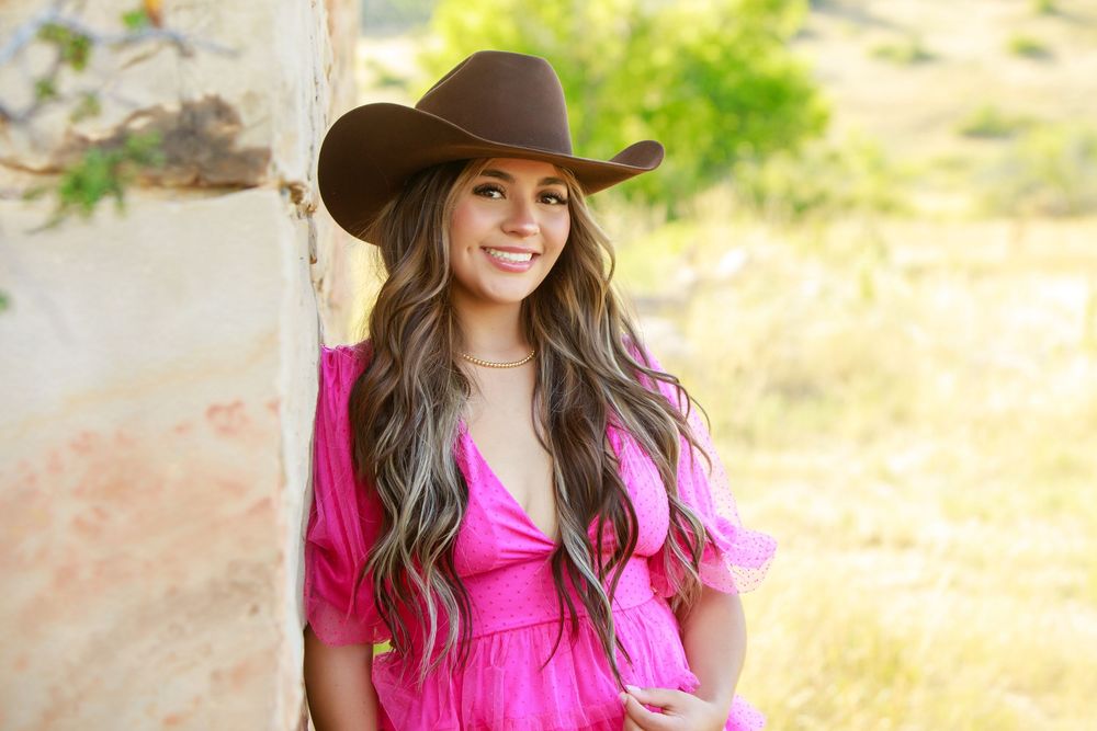 Woman wearing pink dress and brown hat smiles while standing in sunny outdoor setting.