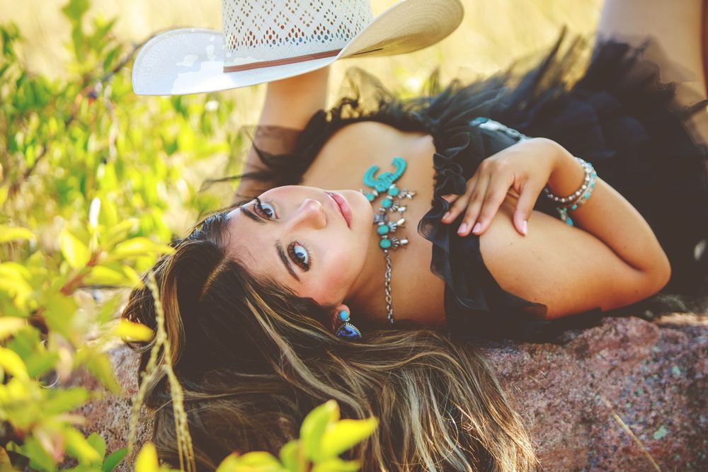 A dreamy outdoor portrait with a white cowboy hat and turquoise jewelry amid green foliage in warm sunlight.