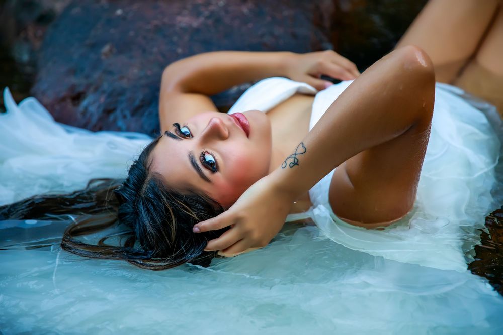 A glamour portrait of a person in water wearing a white garment with dark flowing hair and dramatic makeup.